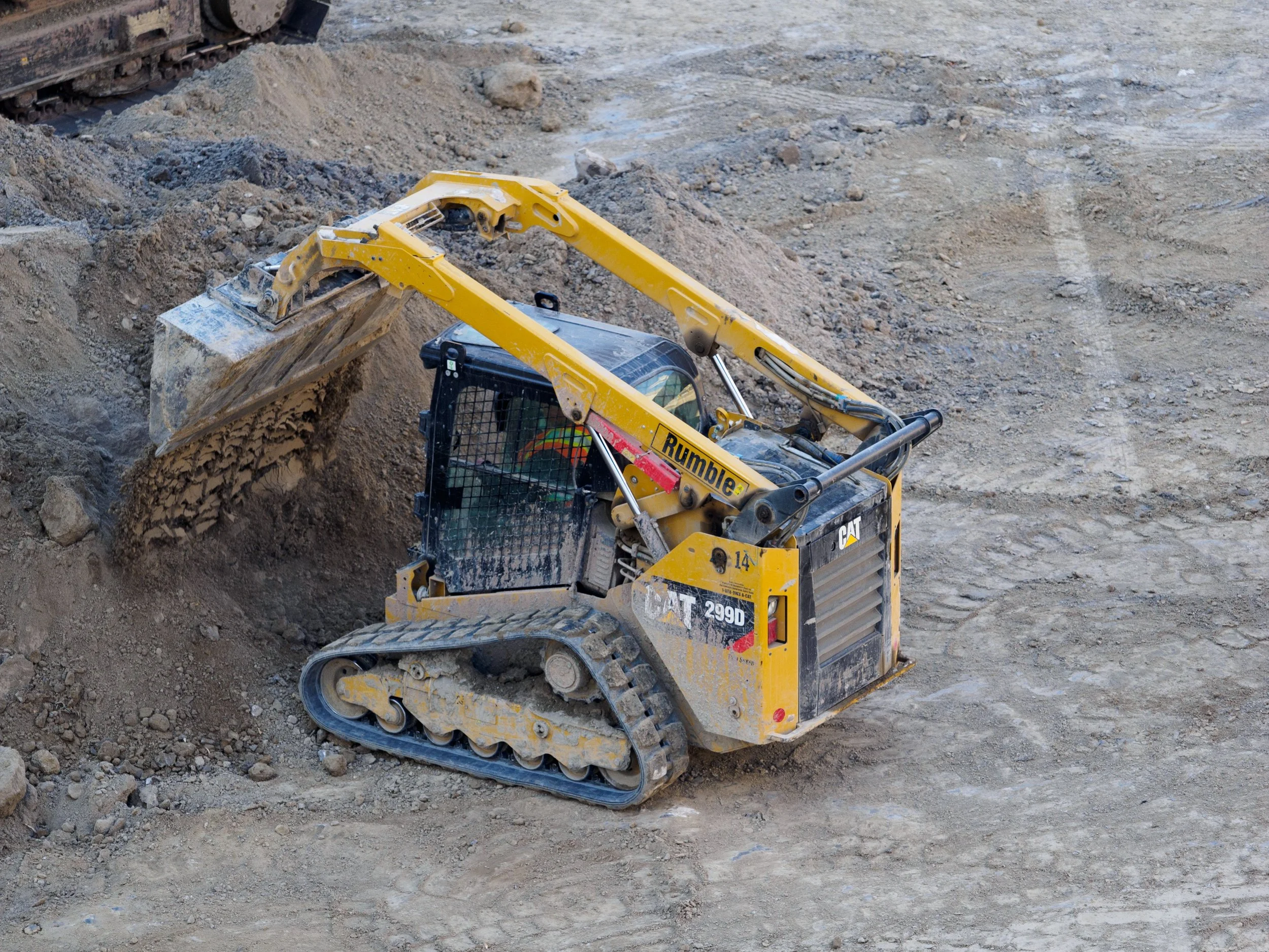 A yellow excavator working on a construction site, digging into the dark soil with rocks scattered around.