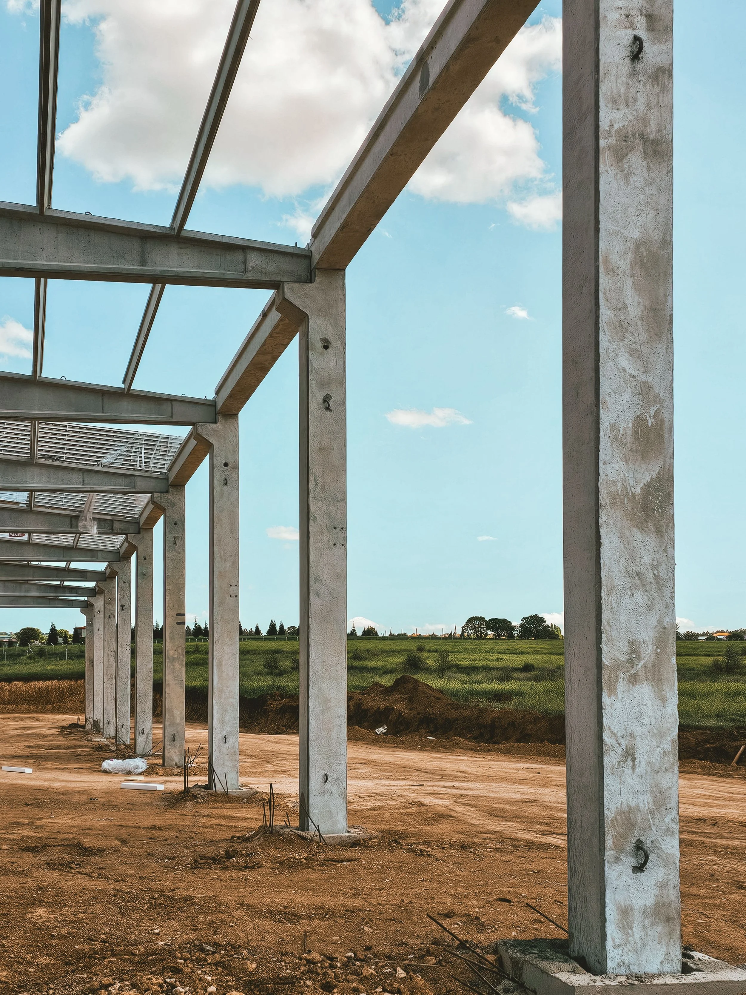 Concrete building framework under construction with vertical supports and open roof, outdoor setting with green fields and trees in the background, blue sky with clouds.