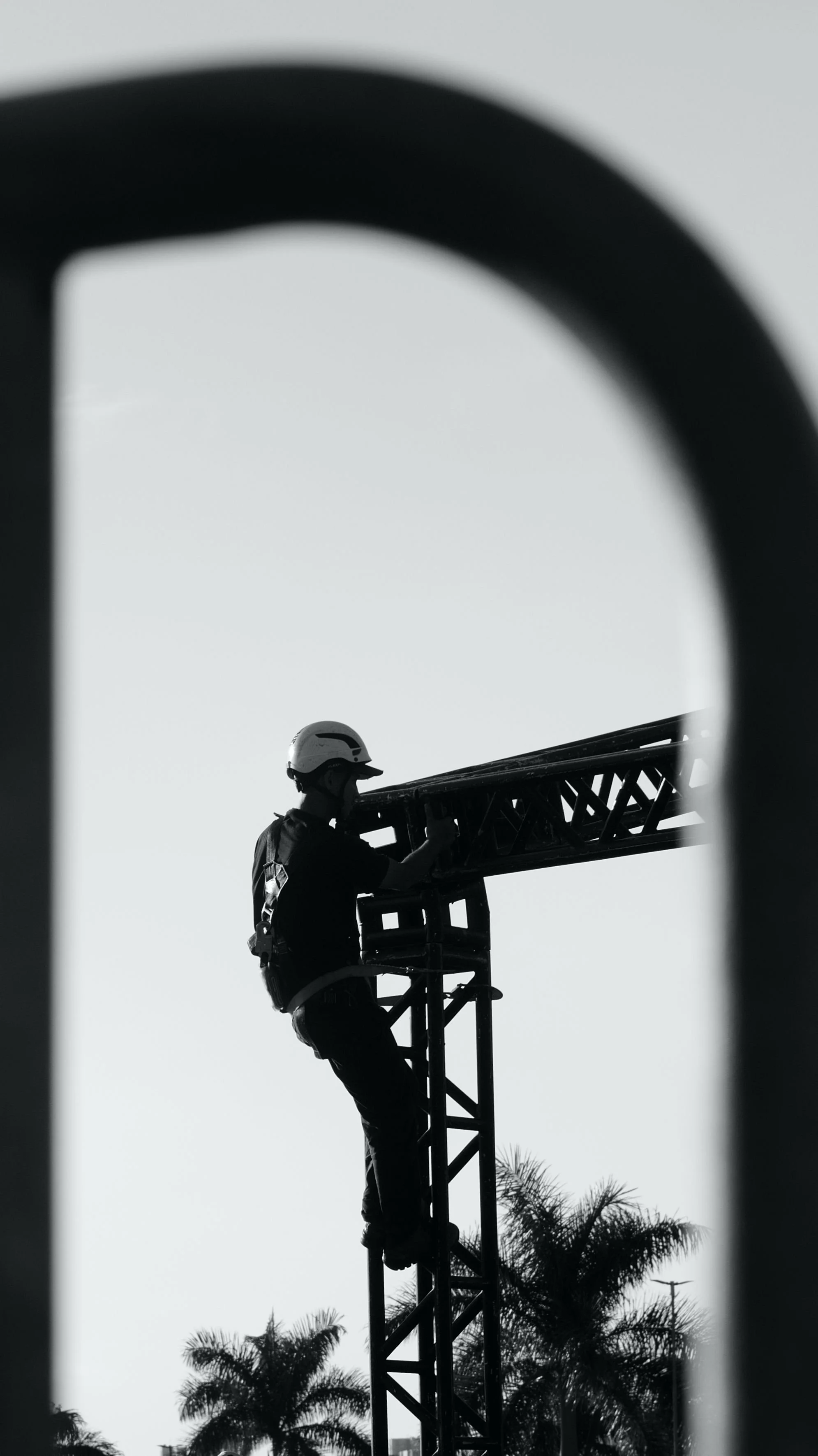 A silhouette of a worker wearing a safety helmet sitting on a steel structure, possibly a radio tower or crane, with trees in the background, viewed through a blurred frame.