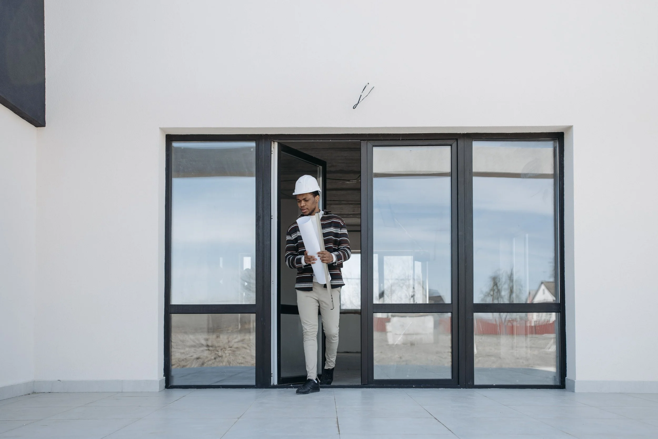 A man in a striped sweater and beige pants standing in front of glass doors holding rolled-up architectural plans, wearing a white safety helmet, outside a modern building.