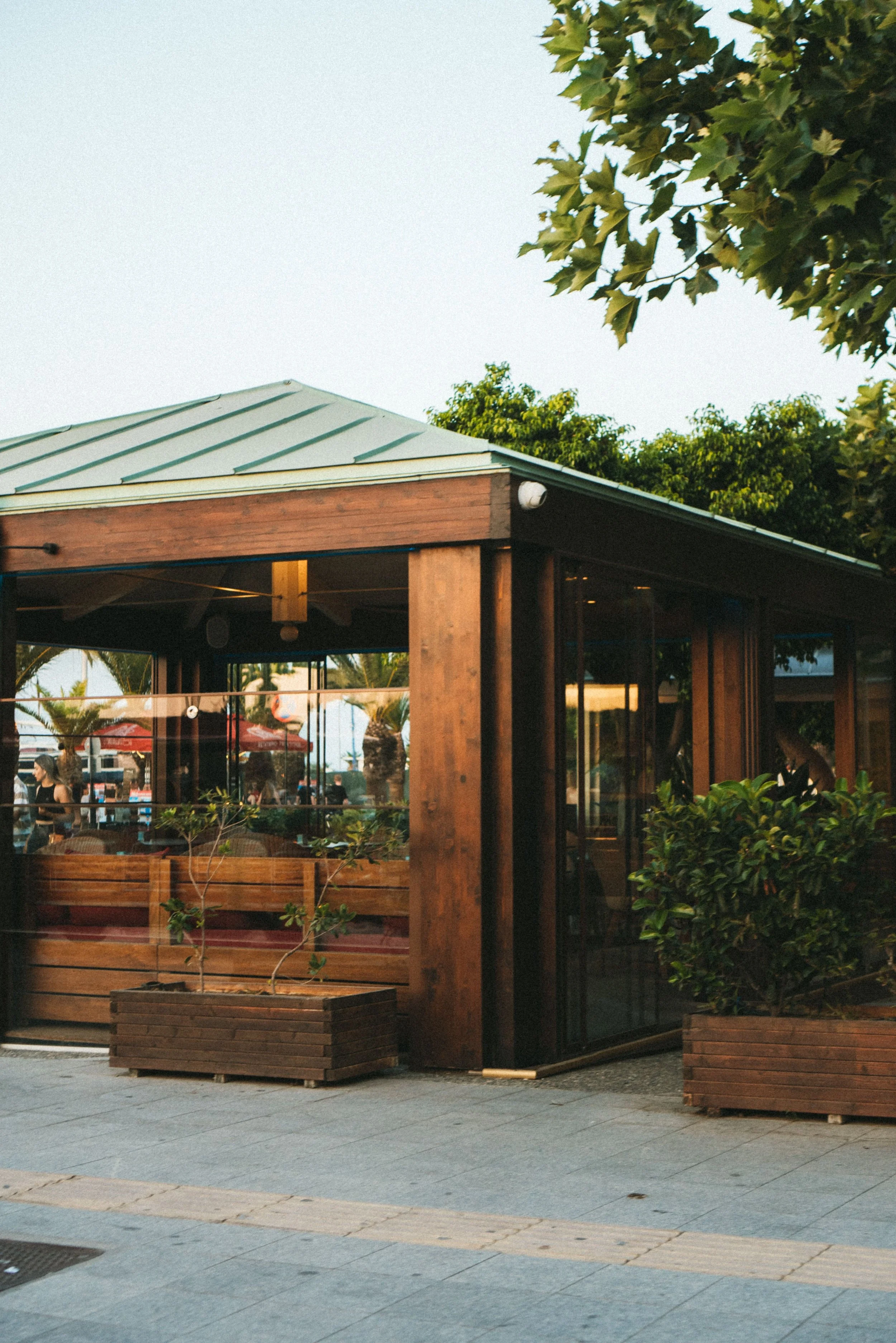 Exterior view of a cozy restaurant or cafe with wooden framing, large windows, and an outdoor patio area with plants in planters, situated in a lively area with outdoor seating and umbrellas visible in the background.