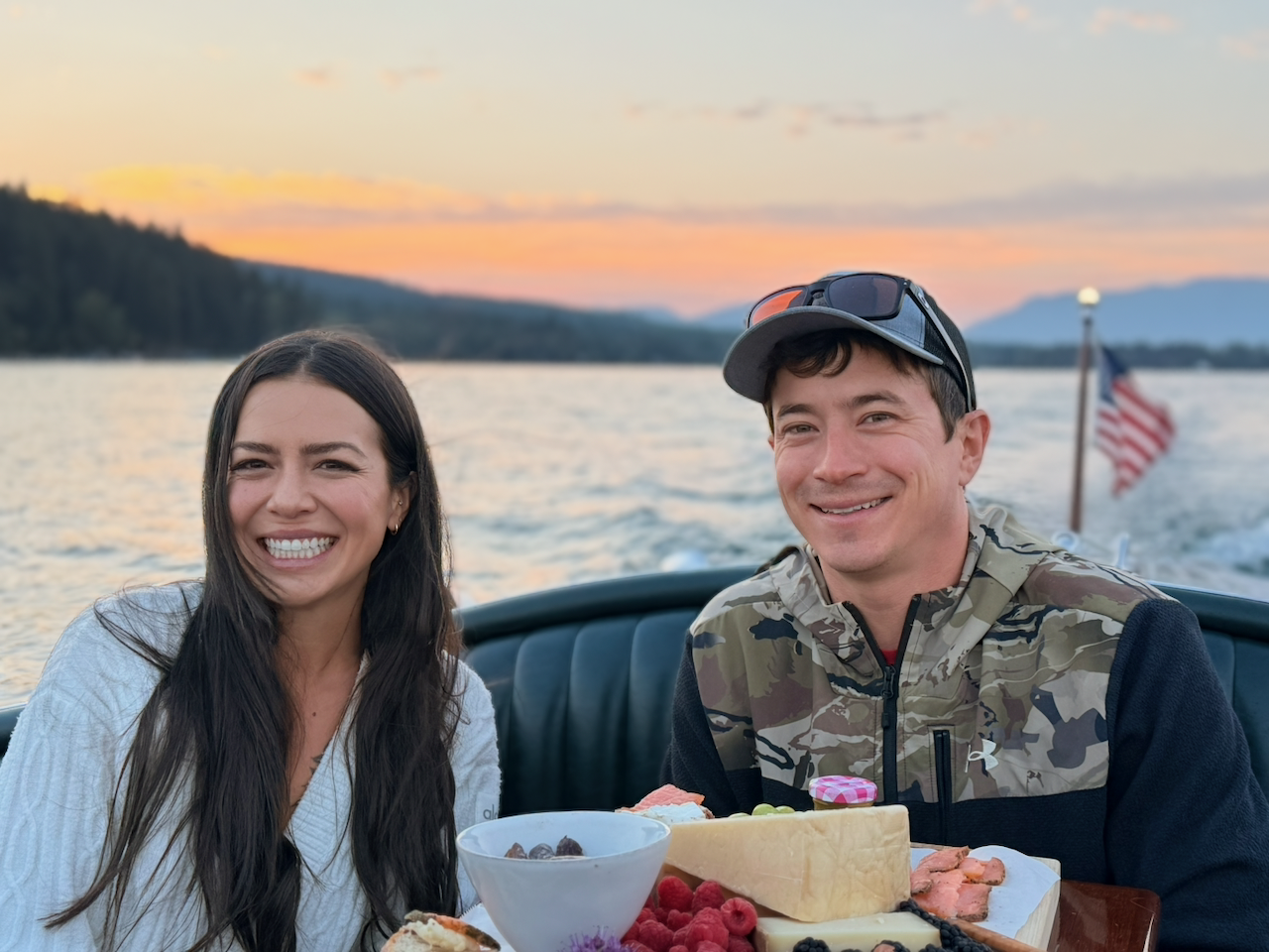 A romantic couple enjoying an early morning 6:30 AM sunrise cruise, with coffee and a breakfast charcuterie board, on the calm waters of Whitefish Lake, Montana.