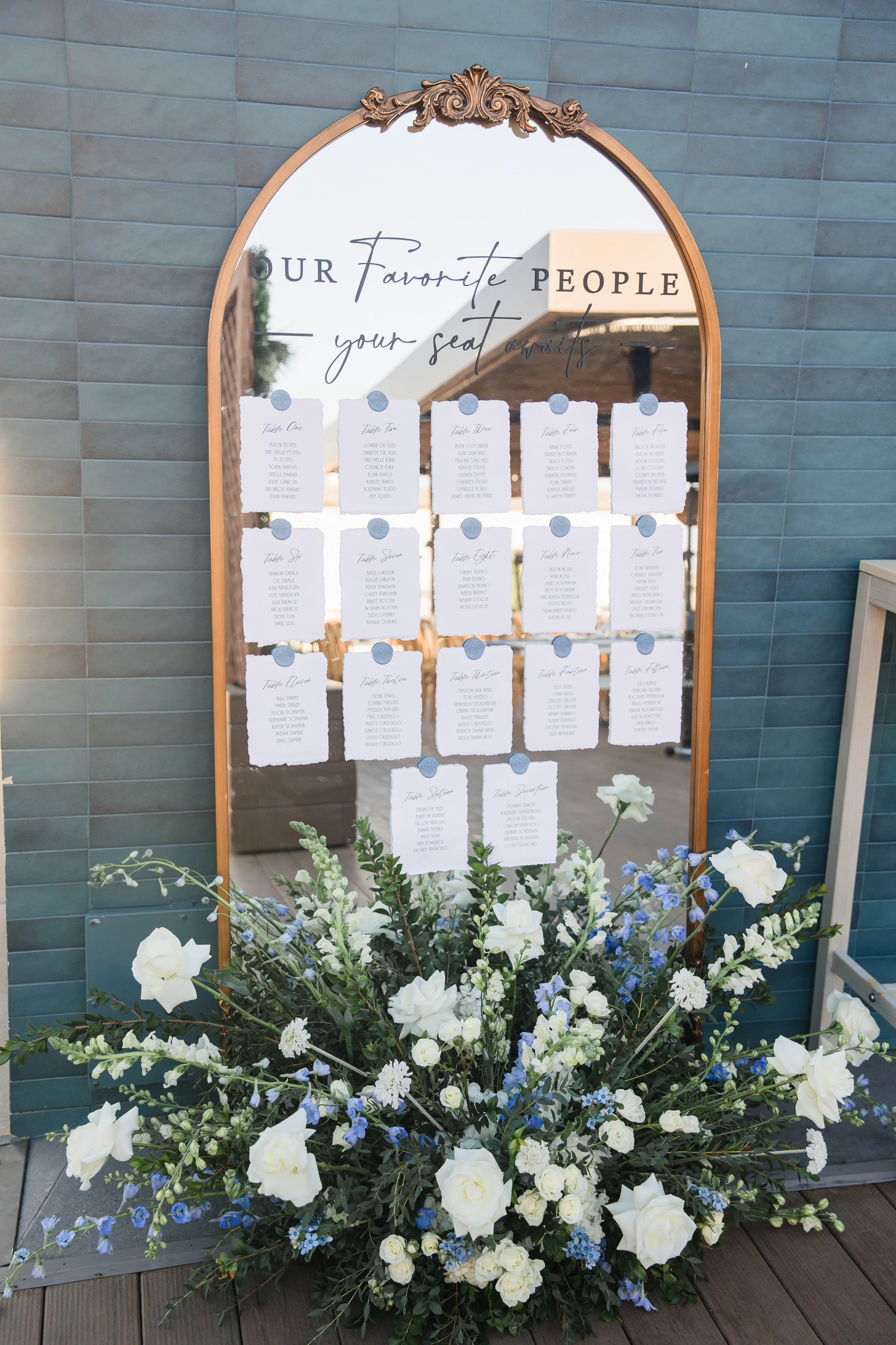 A large floral arrangement with white and blue flowers in front of a mirror with a wooden frame. The mirror has a decorative top and displays a seating chart for an event, with names written on white cards. The background includes a blue tiled wall and a wooden floor, suggesting an outdoor or semi-outdoor setting.