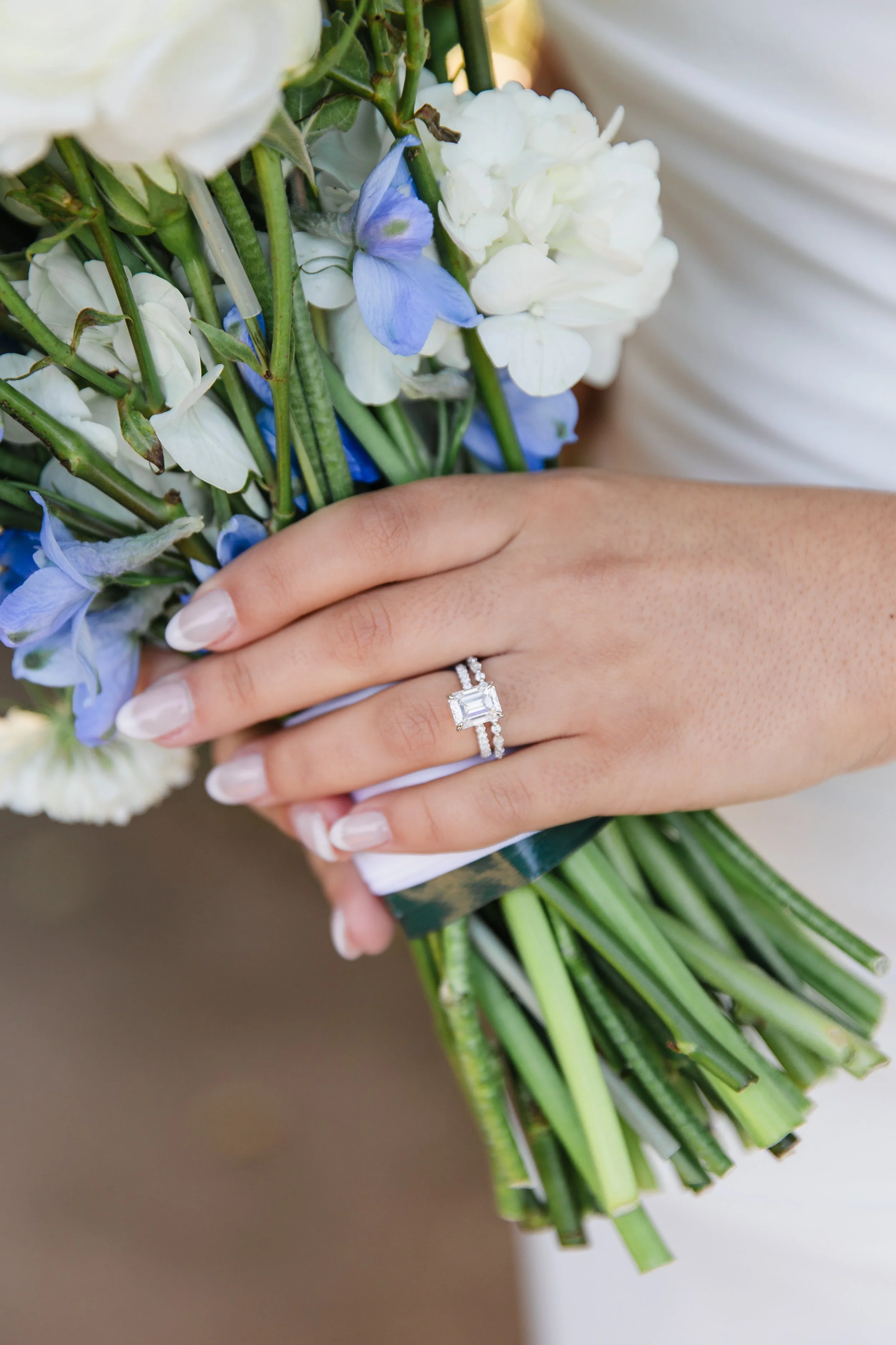 Close-up of a hand holding a bouquet of flowers with white and blue blossoms, wearing a wedding ring with a large square diamond, and a white dress in the background.