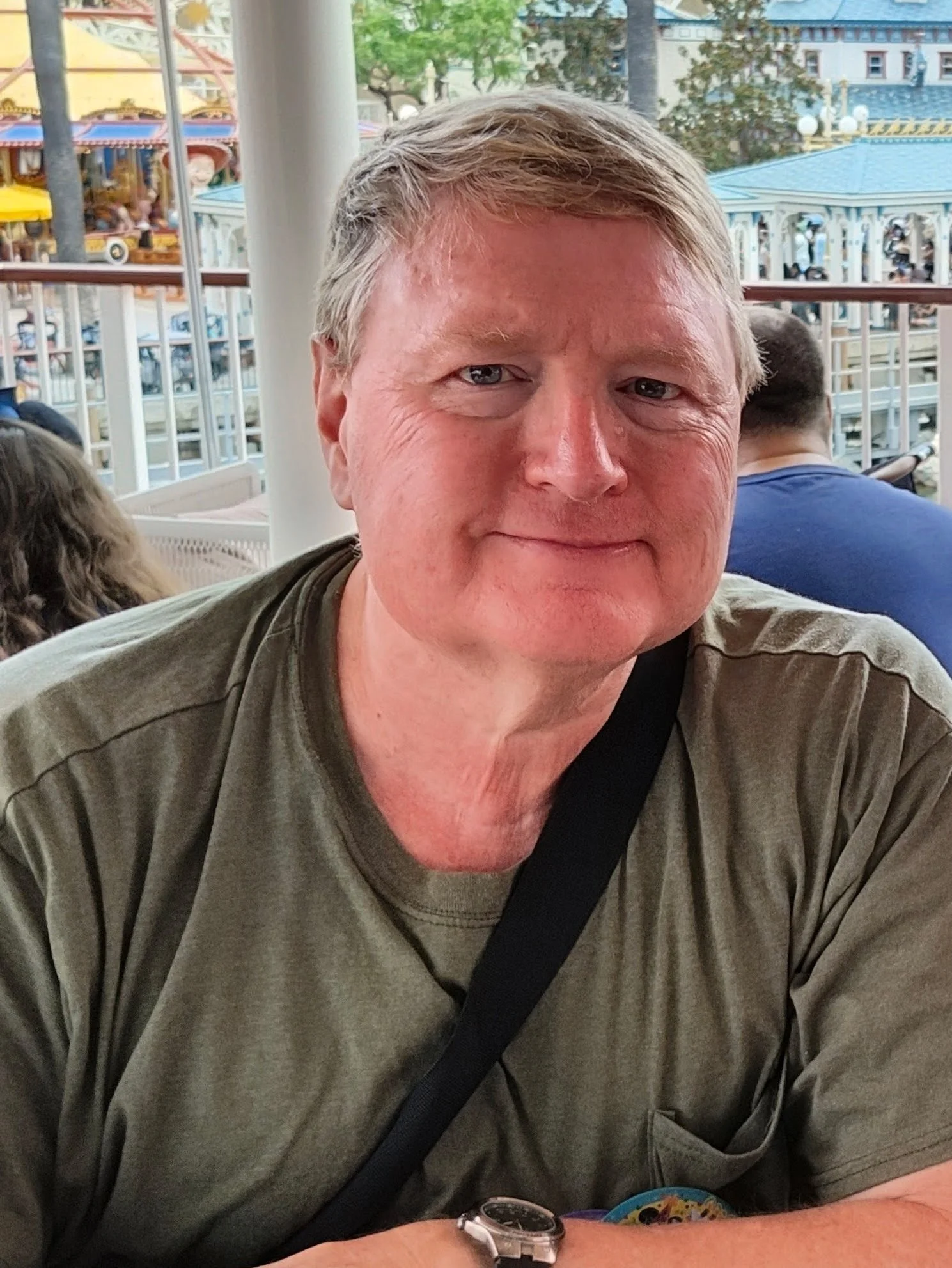 tracysbeck.Close-up of a middle-aged man with light brown hair and blue eyes, smiling, wearing a green shirt with a black strap across his chest, and a watch on his left wrist. The background shows a colorful amusement park with a carousel and trees.