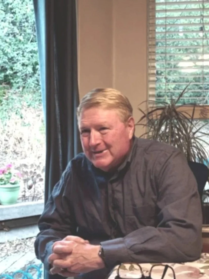 tracysbeck.com. A man with blonde hair sitting at a table near a window with blinds, with a potted plant and outdoor greenery in the background.