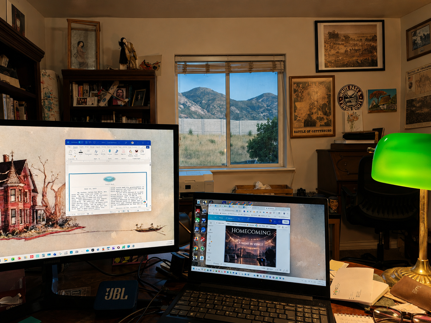 Home office setup with two monitors, a laptop showing a Broadway show, a green desk lamp, a window with mountains in the background, and various framed pictures and posters on the walls.
