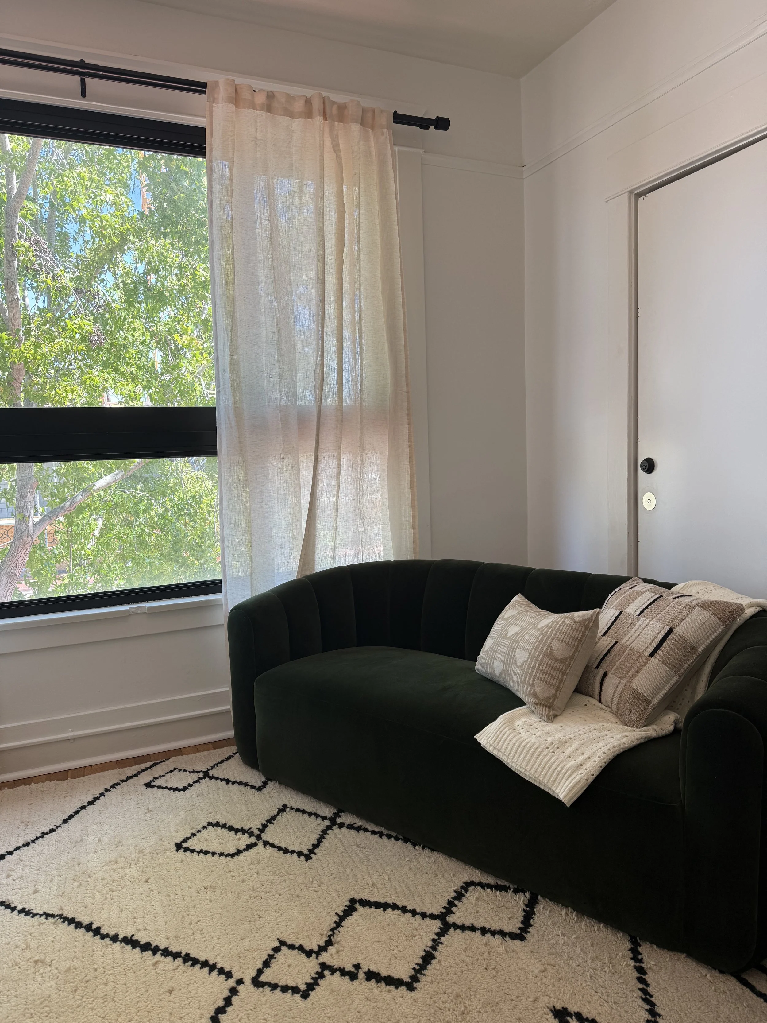 Sunlight streaming through a window in an inviting therapy office with sheer white curtains, dark green sofa with pillows, white textured throw blanket, beige and brown pillows, white and black patterned rug.