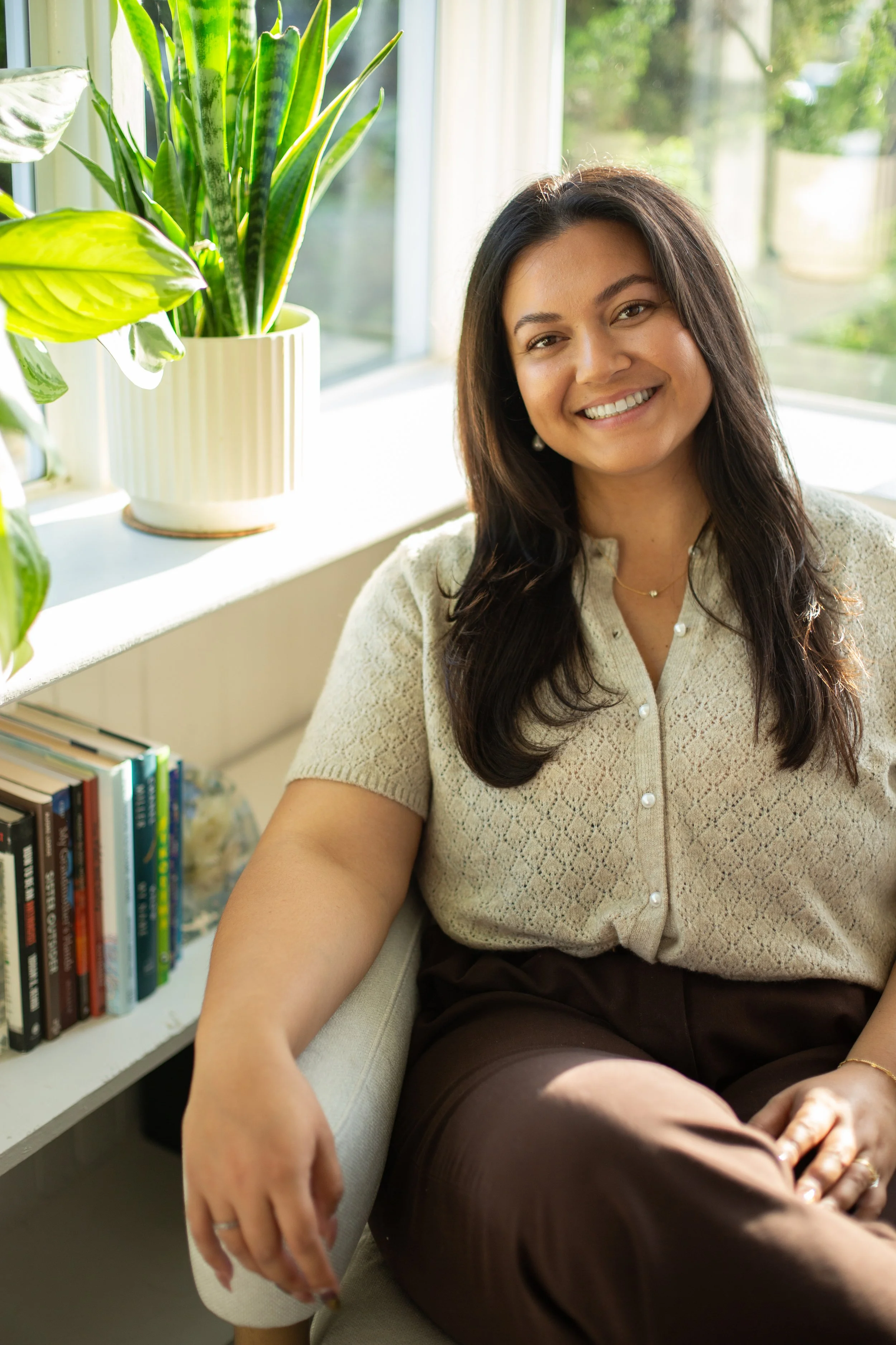 Marisa Dao, Licensed Professional Clinical Counselor, smiling warmly in her cozy San Diego office, while sitting near a window with sunlight, next to a green potted plant and a bookshelf with therapy books.