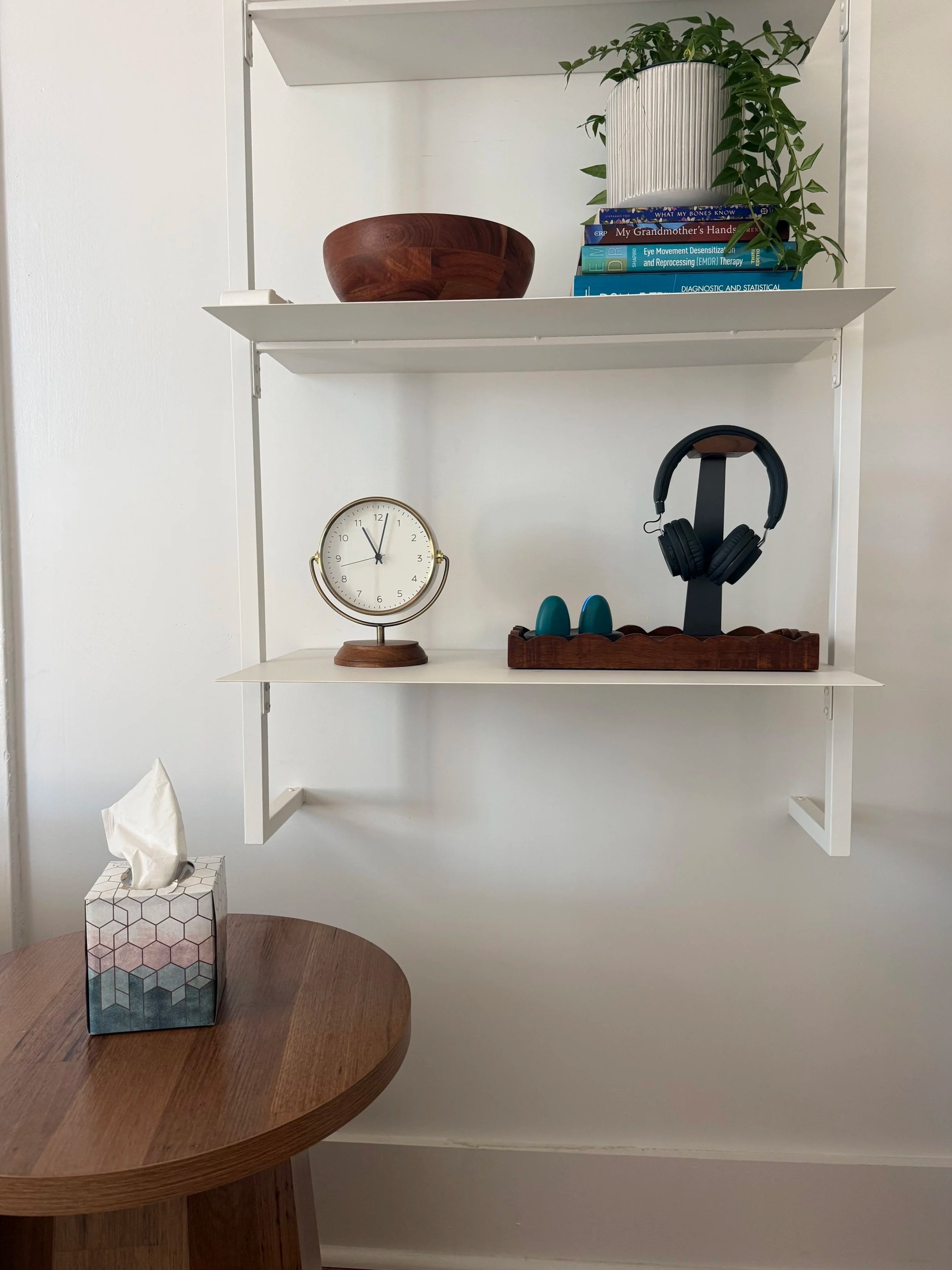 A white shelf in a therapy office with a wooden bowl with sour candy/mints, a stack of therapy books, and a potted plant. Below the shelf is a clock and a stand with EMDR headphones and EMDR pulsators. In the foreground is a table with a tissue box.