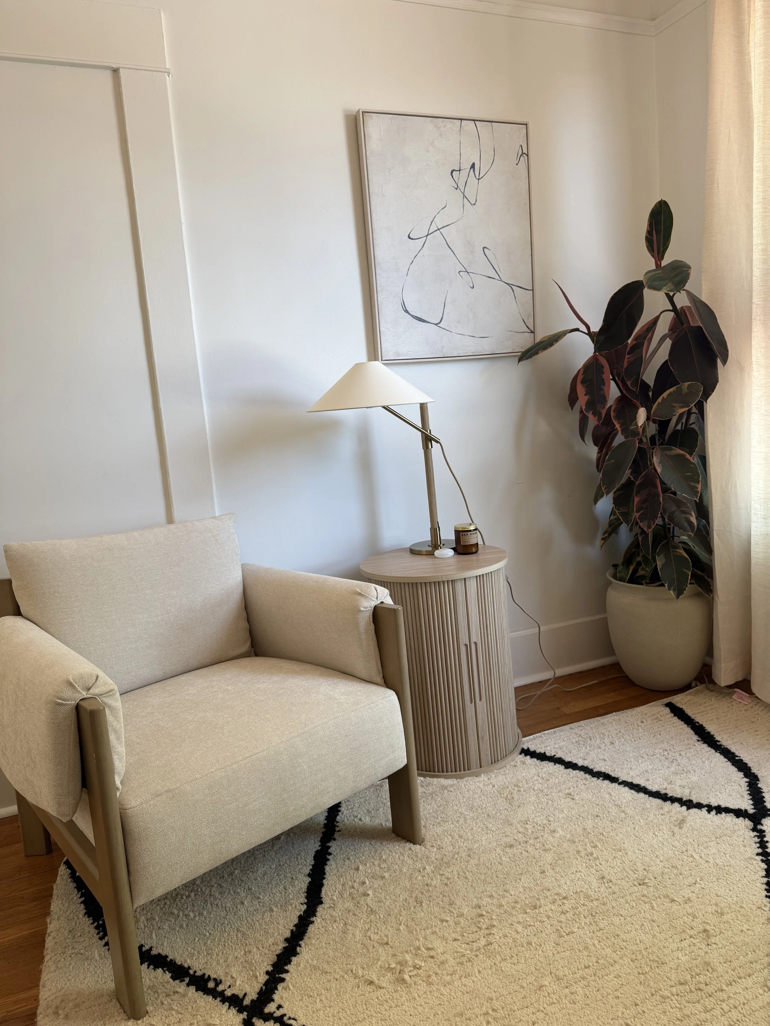 A cozy therapy office corner with a beige therapist armchair, a small wooden side table with a modern brass lamp and candle, a large potted plant, a textured white and black rug, and a framed abstract line art on the wall near a large window.