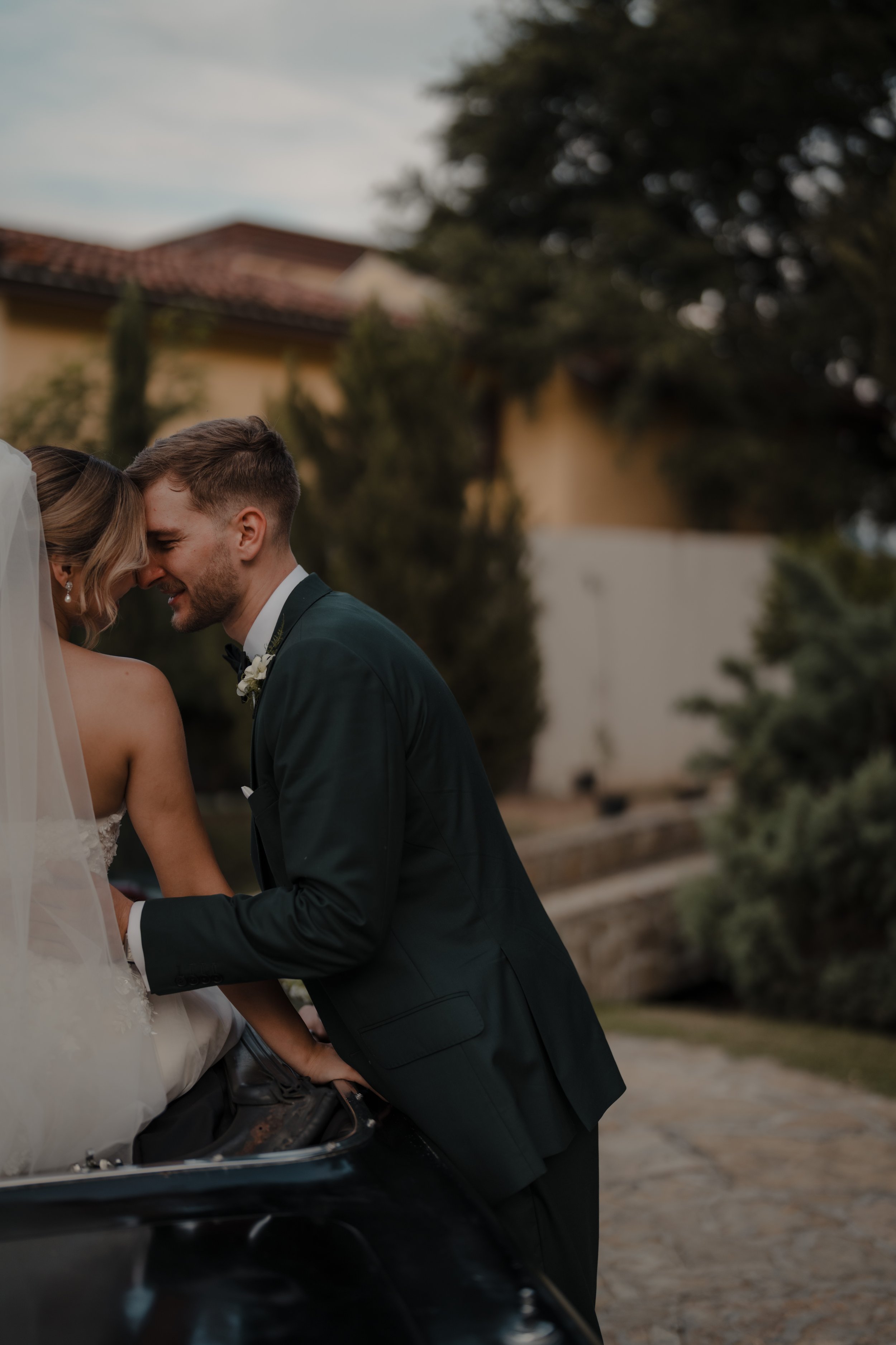 Pareja de recién casados riendo, en traje de boda, en una ceremonia al aire libre, junto a un coche clásico y árboles, en un ambiente de celebración.
