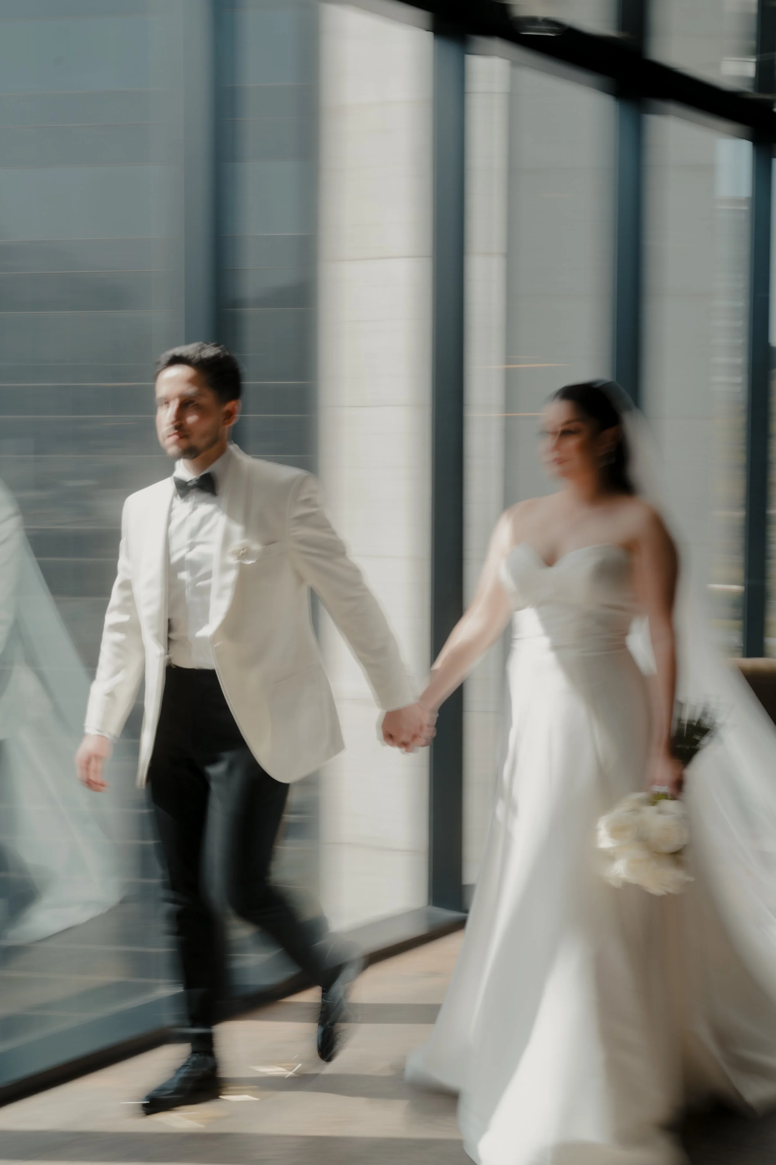 Una pareja en un vestido de novia y un traje de etiqueta caminando tomada a través de un ventanal en un edificio moderno.