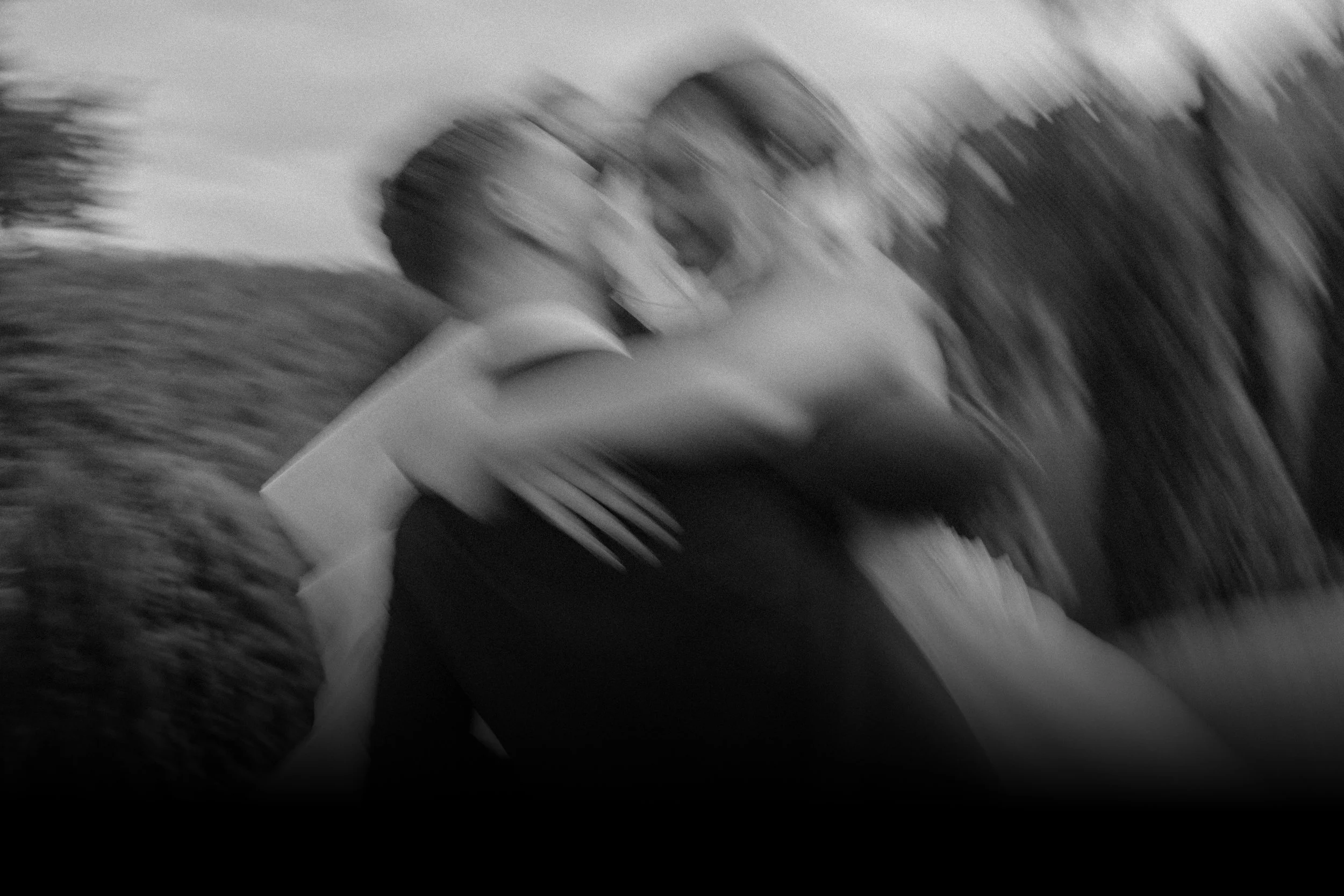 Imagen en blanco y negro de una pareja besándose al aire libre, con árboles y cielo de fondo, con efecto de movimiento