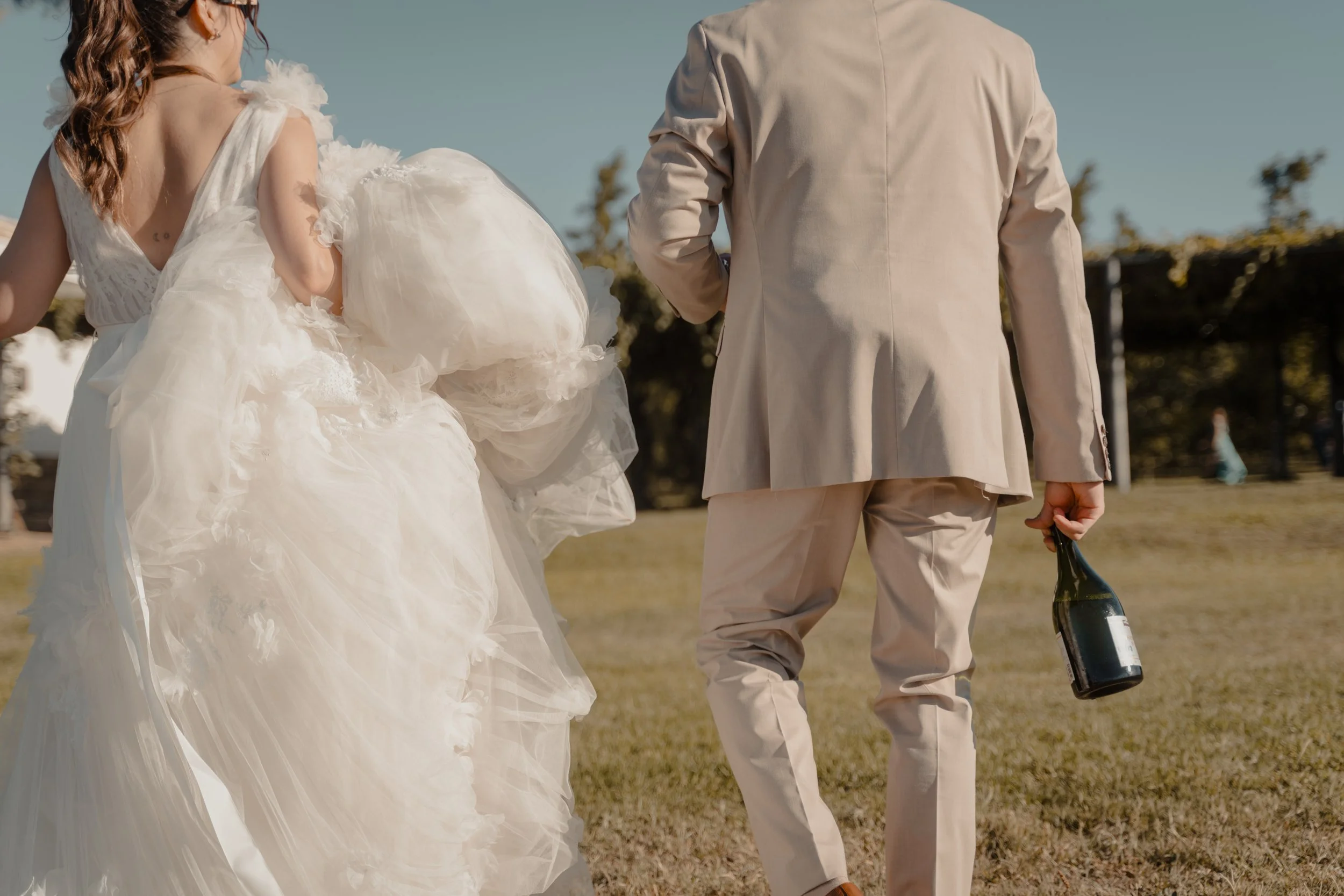Una pareja en una ceremonia de boda al aire libre, la mujer lleva un vestido de novia blanco y el hombre un traje beige, sosteniendo una botella de vino.