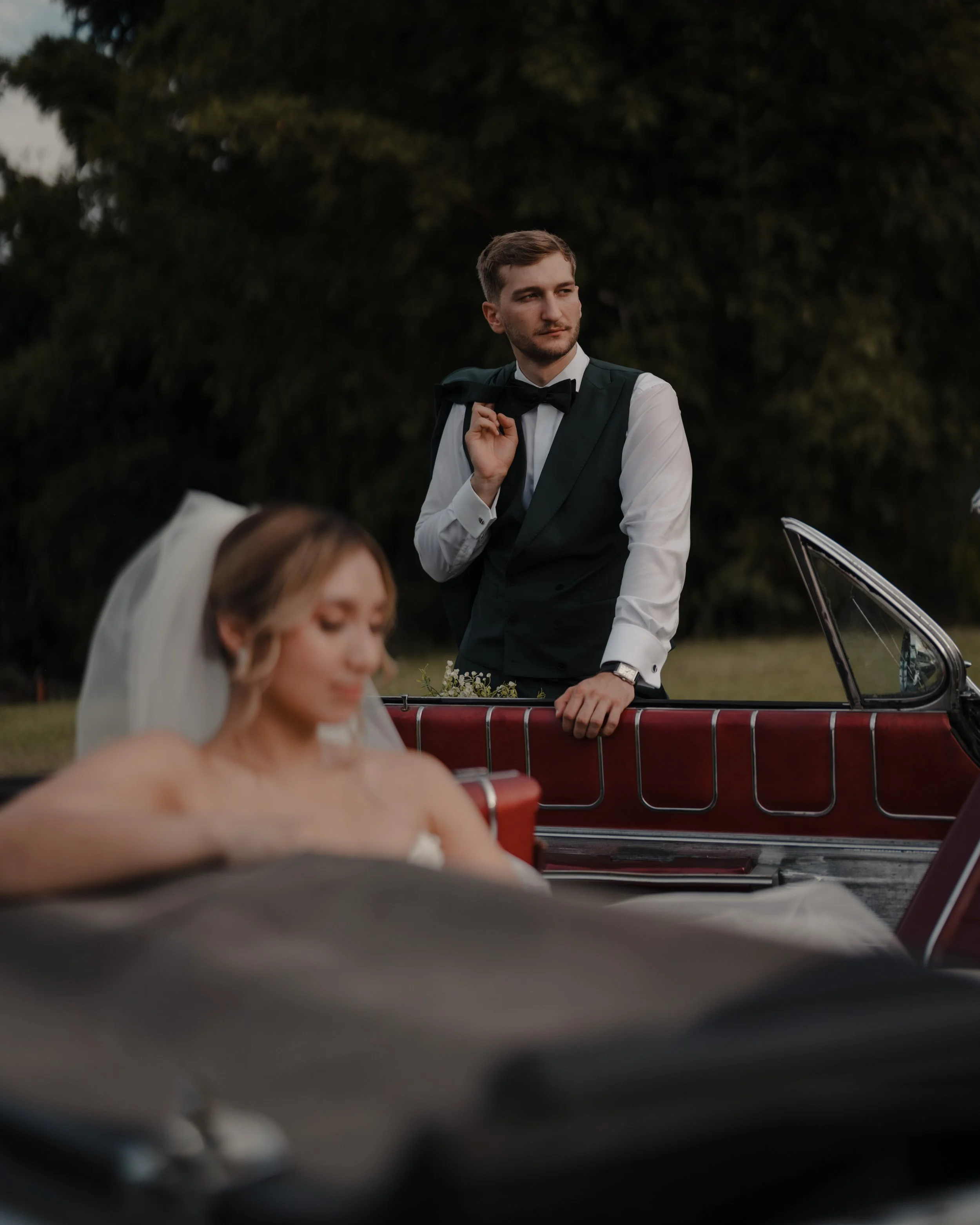 Un hombre vestido con esmoquin, con barba, sosteniendo su saco sobre el hombro, de fondo árboles y un automóvil rojo.