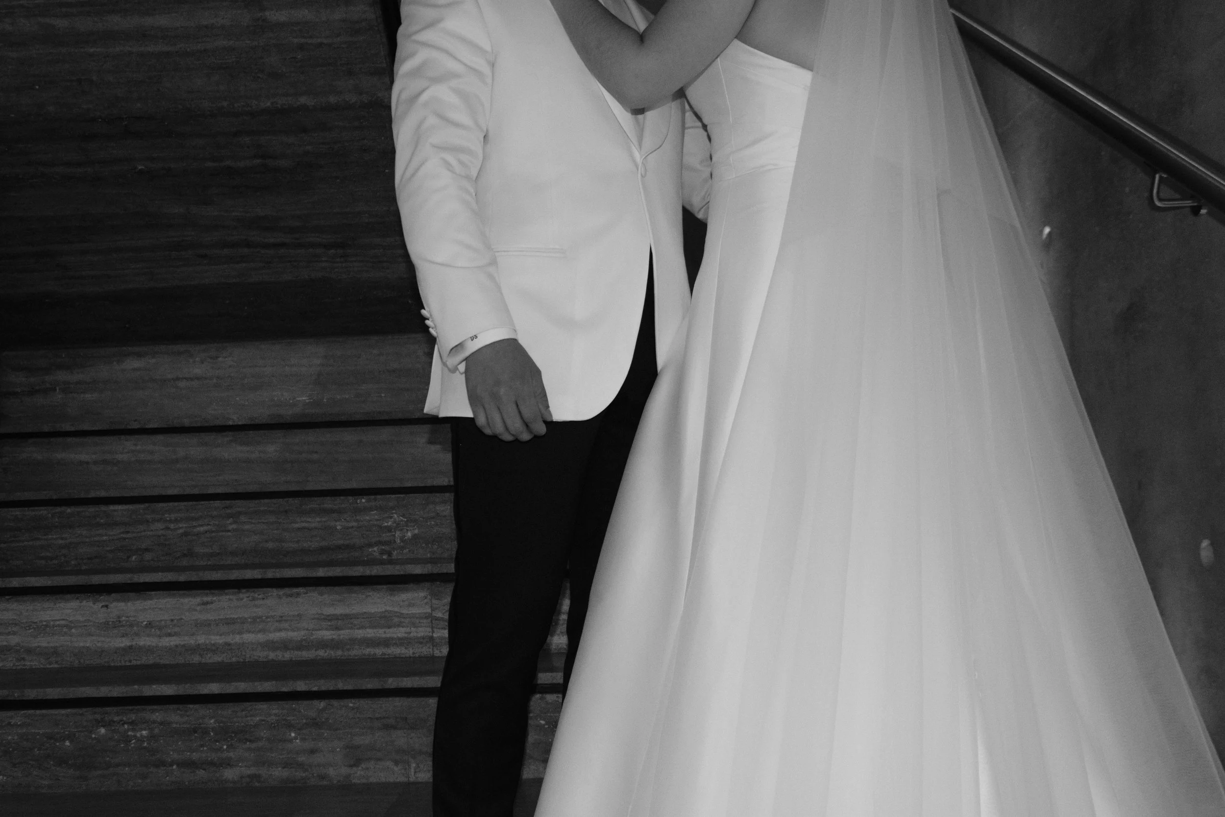 Pareja en vestimenta formal, la mujer lleva vestido de novia y el hombre lleva saco blanco, en un entorno interior con suelo de madera.