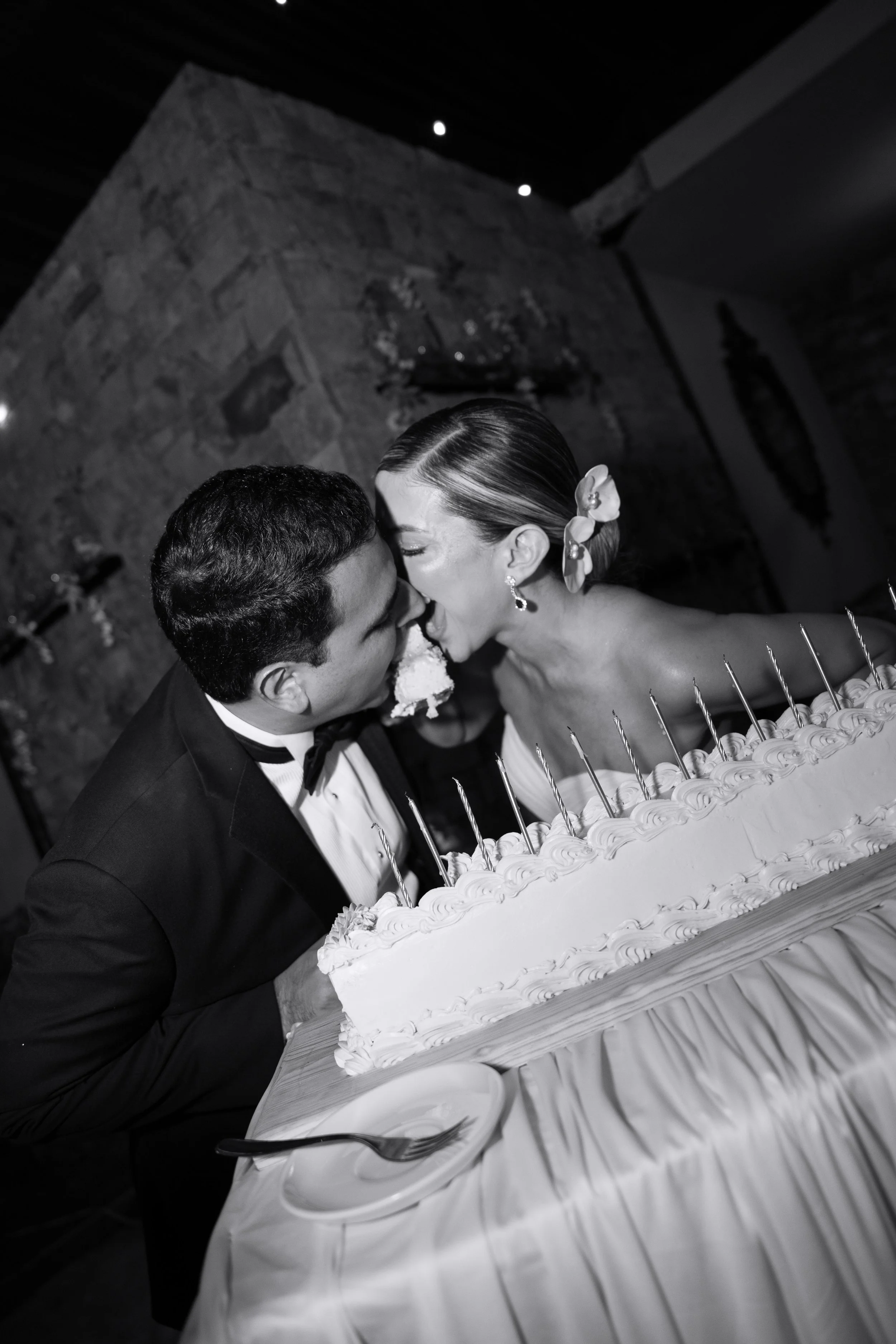 Una pareja vestida formalmente compartiendo un beso frente a un pastel de bodas decorado con crema y decoraciones. La cena y la celebración parecen ser en un lugar con paredes de piedra.