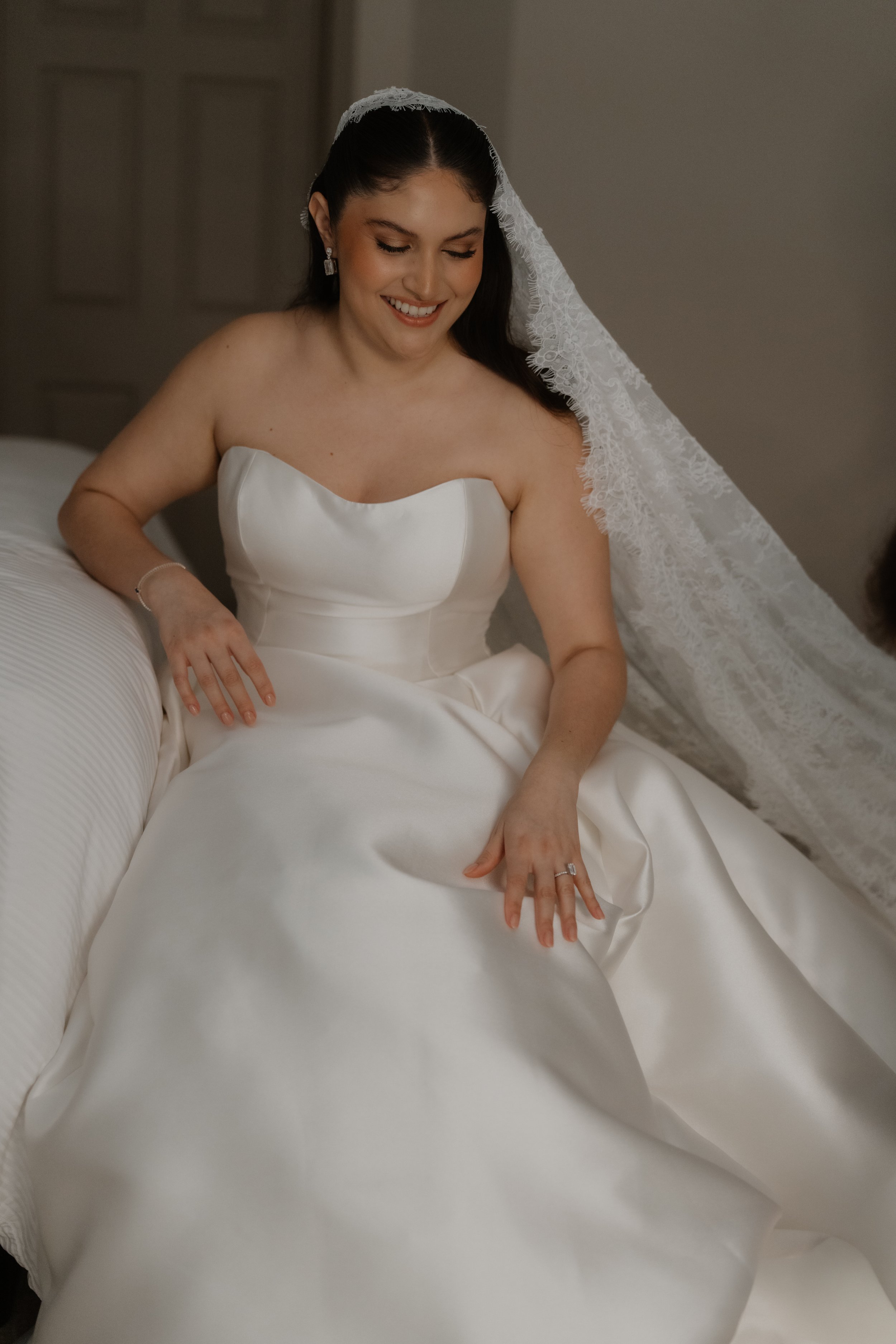 Mujer con vestido de novia y velo, sonriendo, sentada en una cama.