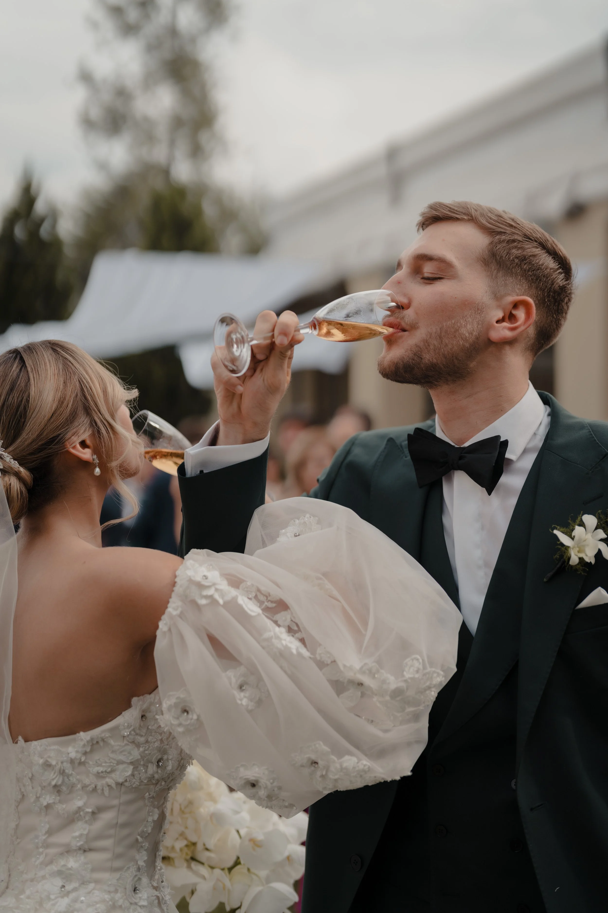 Una pareja de novios bailando en su boda, la novia lleva un vestido blanco con detalles de encaje y la pareja está brindando con copa de vino rosado, en un ambiente de celebración exterior.