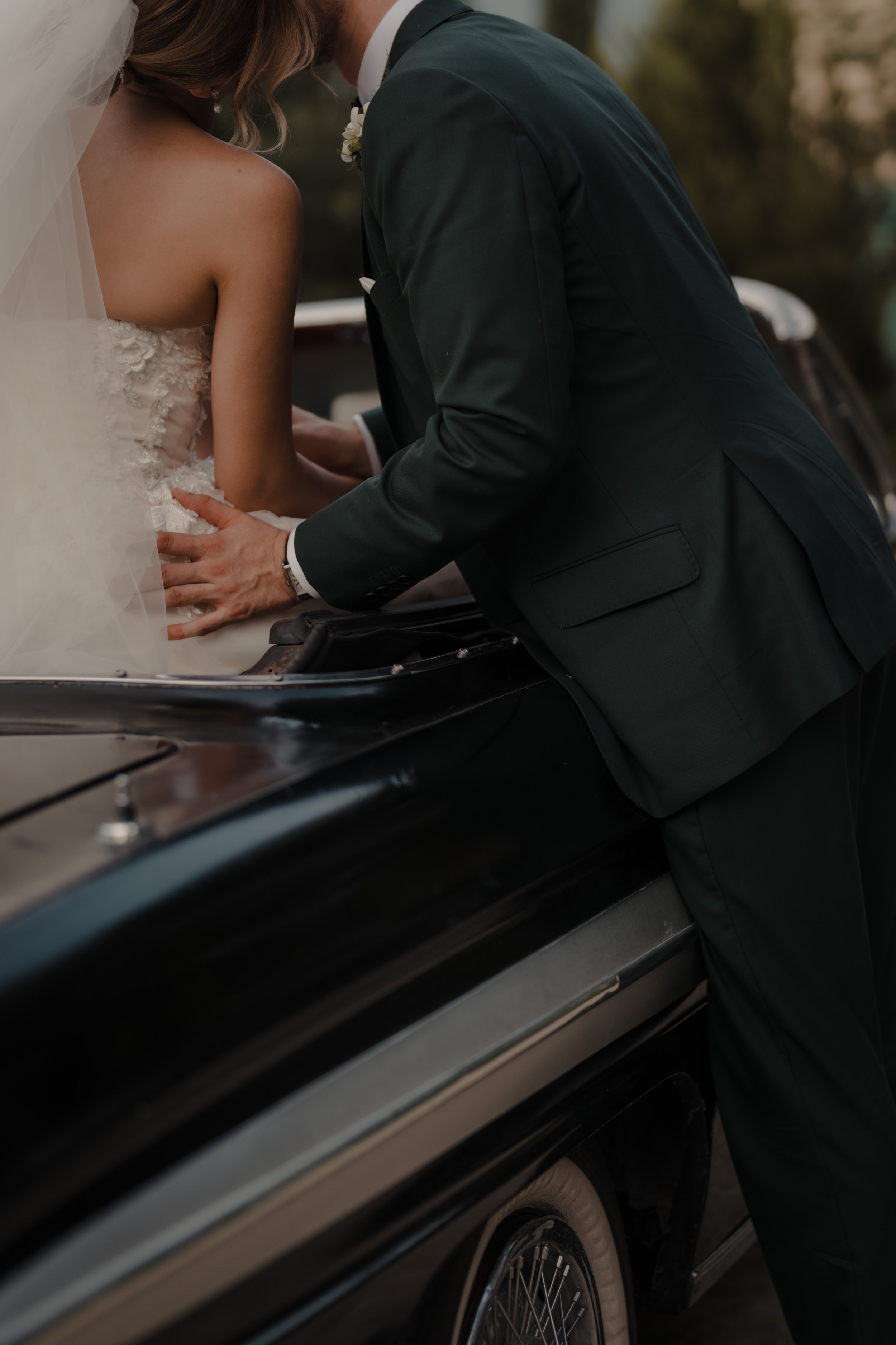 Un hombre y una mujer en vestido de novia y traje formal, besándose en la capota de un coche clásico durante su boda, en un entorno al aire libre con árboles.