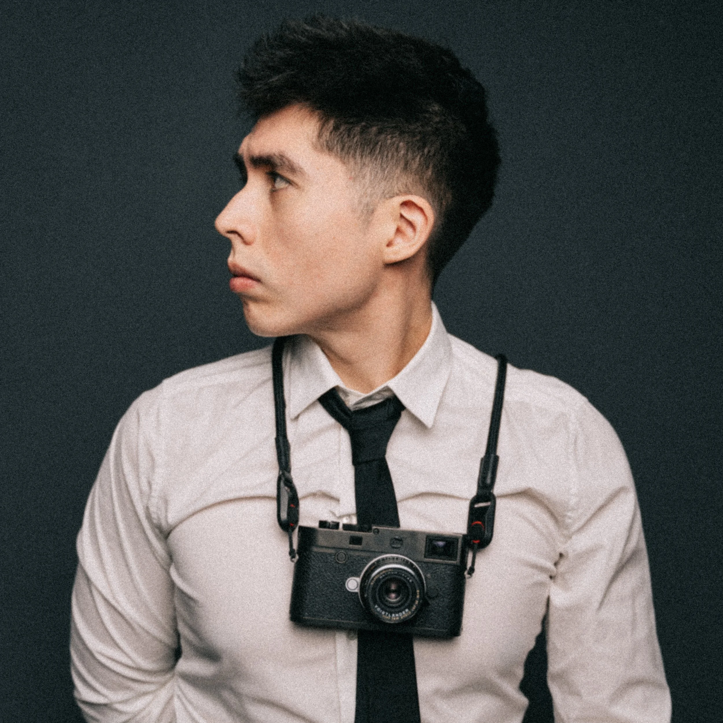 Joven con cabello corto y oscuro, vestido con camisa blanca y chaqueta negra, con una cámara fotográfica alrededor del cuello, apoyada en su pecho, en un fondo oscuro.