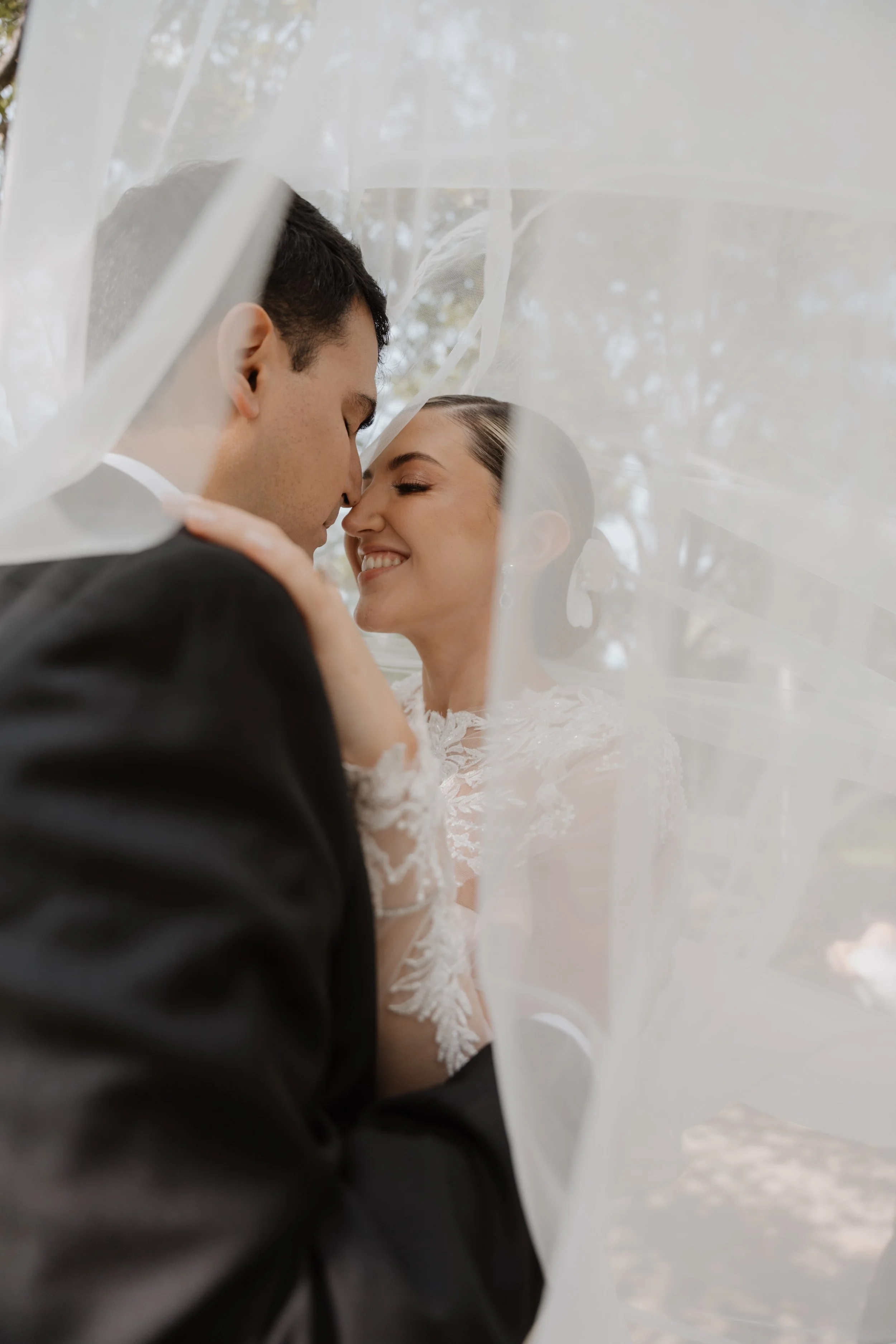 Pareja de recién casados besándose bajo un arco de velo de novia. La mujer sonrie feliz y el hombre la sostiene con ternura.