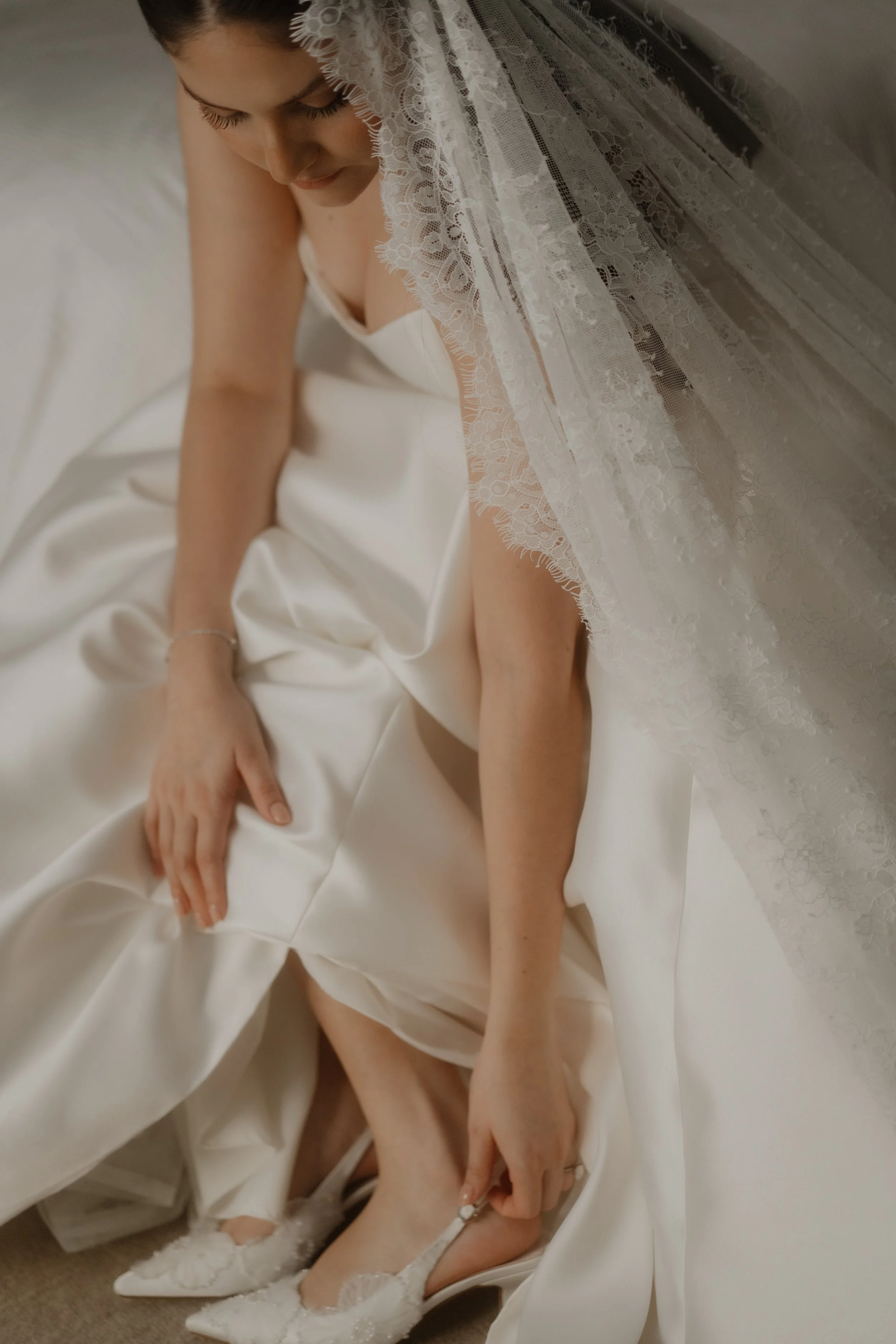 Una mujer con vestido blanco, con velo de encaje, se está poniendo tacones blancos con detalles de encaje y pedrería, en un ambiente interior con alfombra beige.
