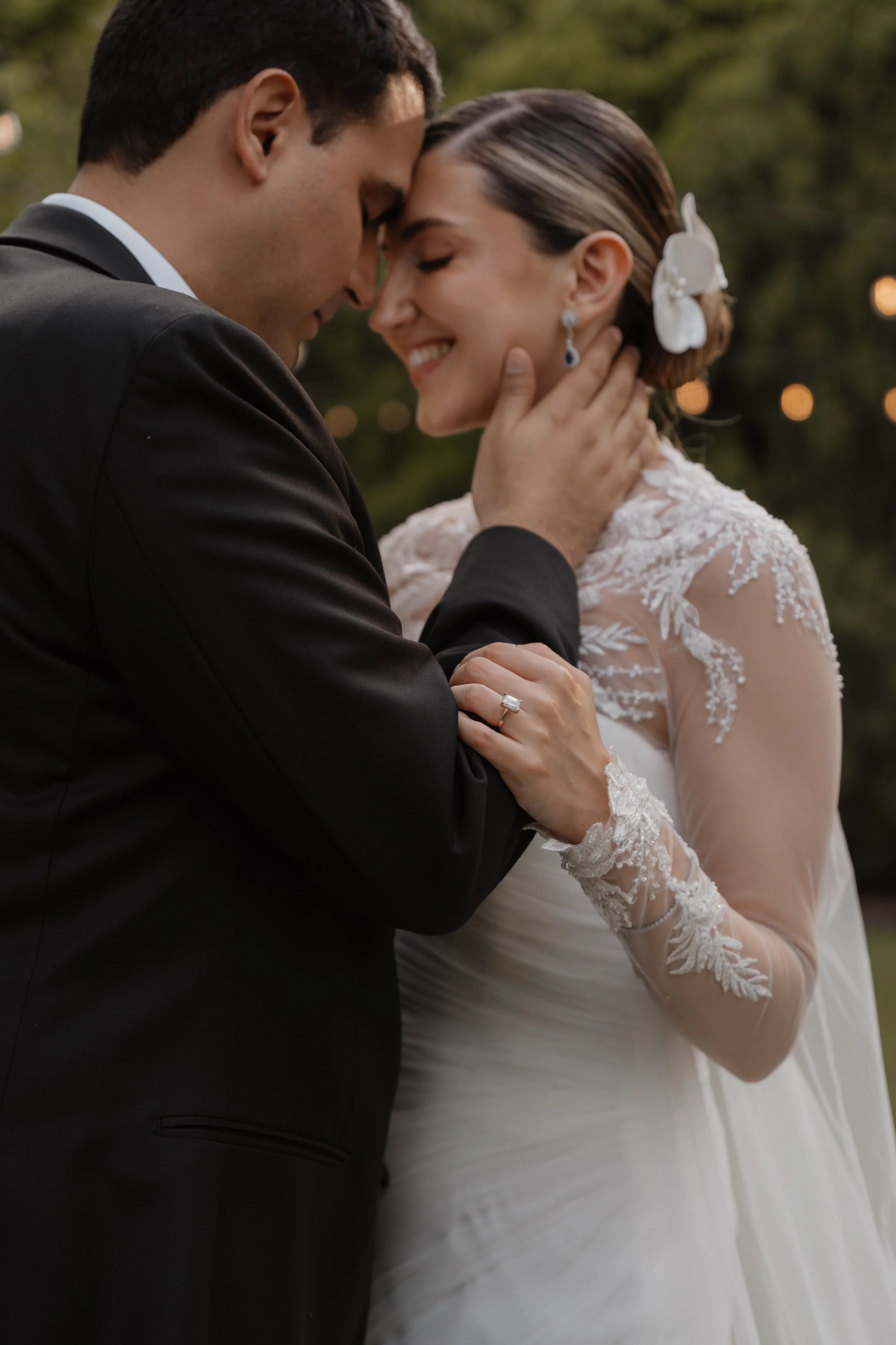 Una pareja en su boda, el hombre vestido con traje negro y la mujer en un vestido de novia con encaje, en un abrazo cariñoso con los ojos cerrados, en un fondo de árboles y luces difusas.