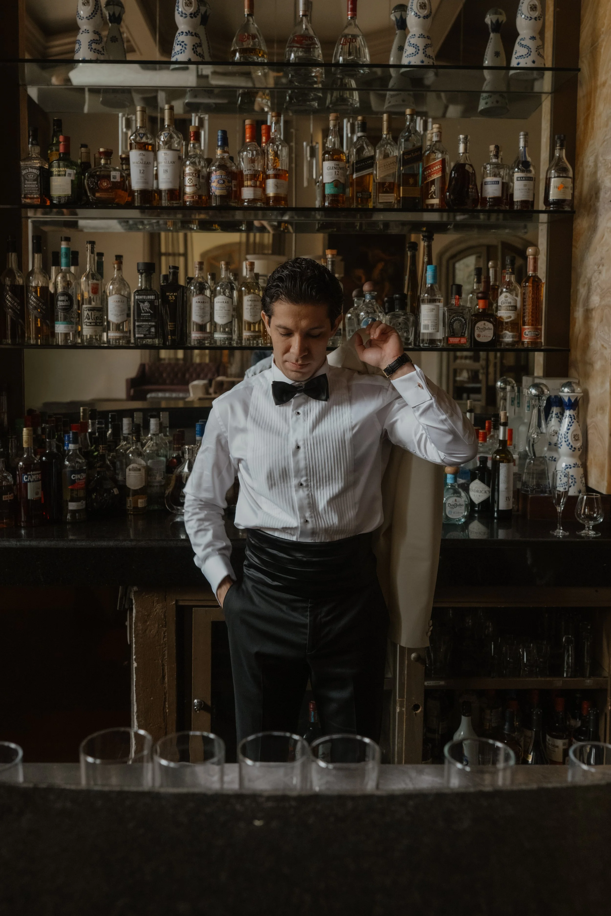 Un bartender con pajarita y peinado peinado, de camisa blanca, en un bar con estantes llenos de botellas de licor.