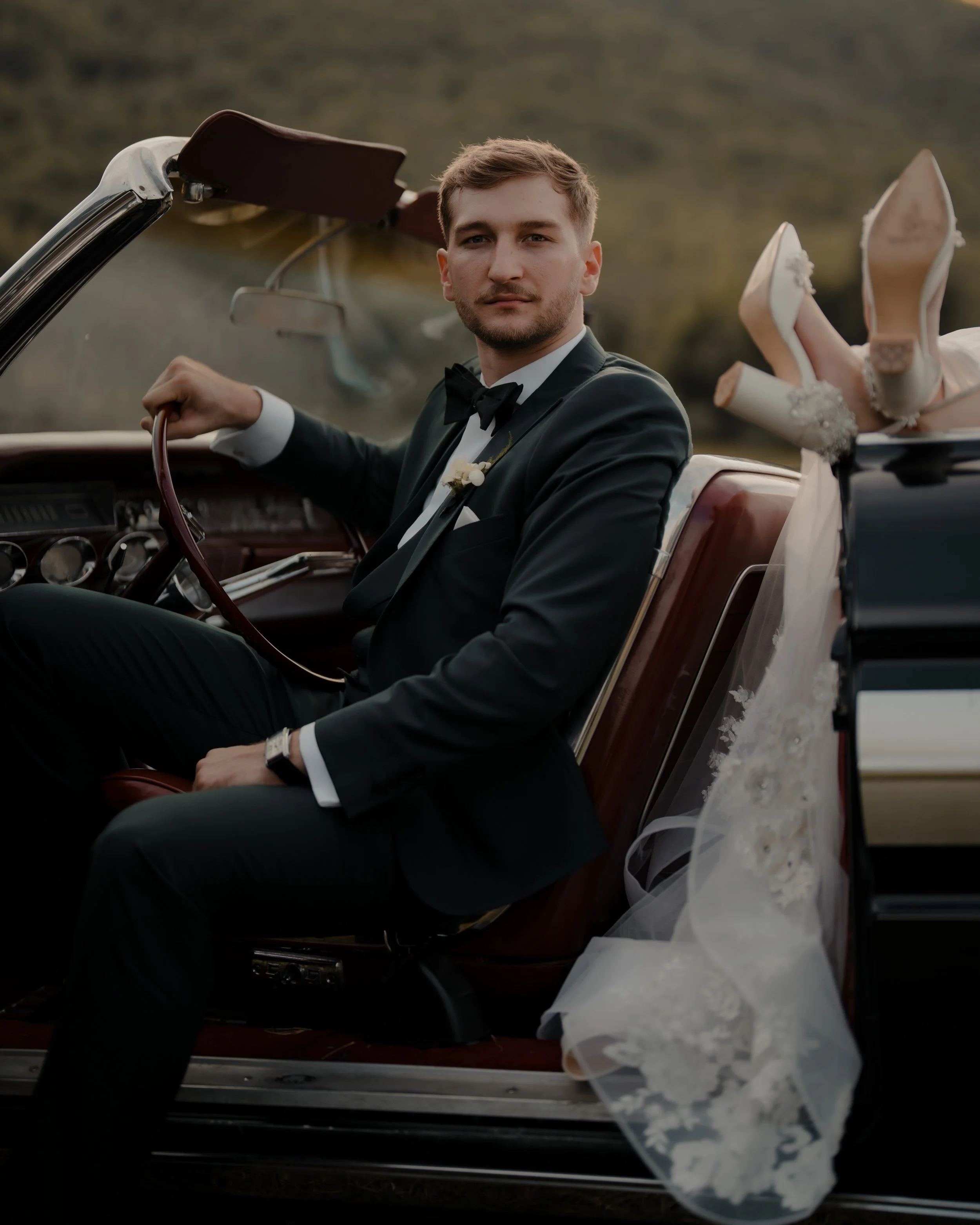 Hombre joven vestido con traje de novio en un coche convertible, con una novia que lleva un vestido de novia con velo y tacones, en un paisaje de campo en tonos cálidos.