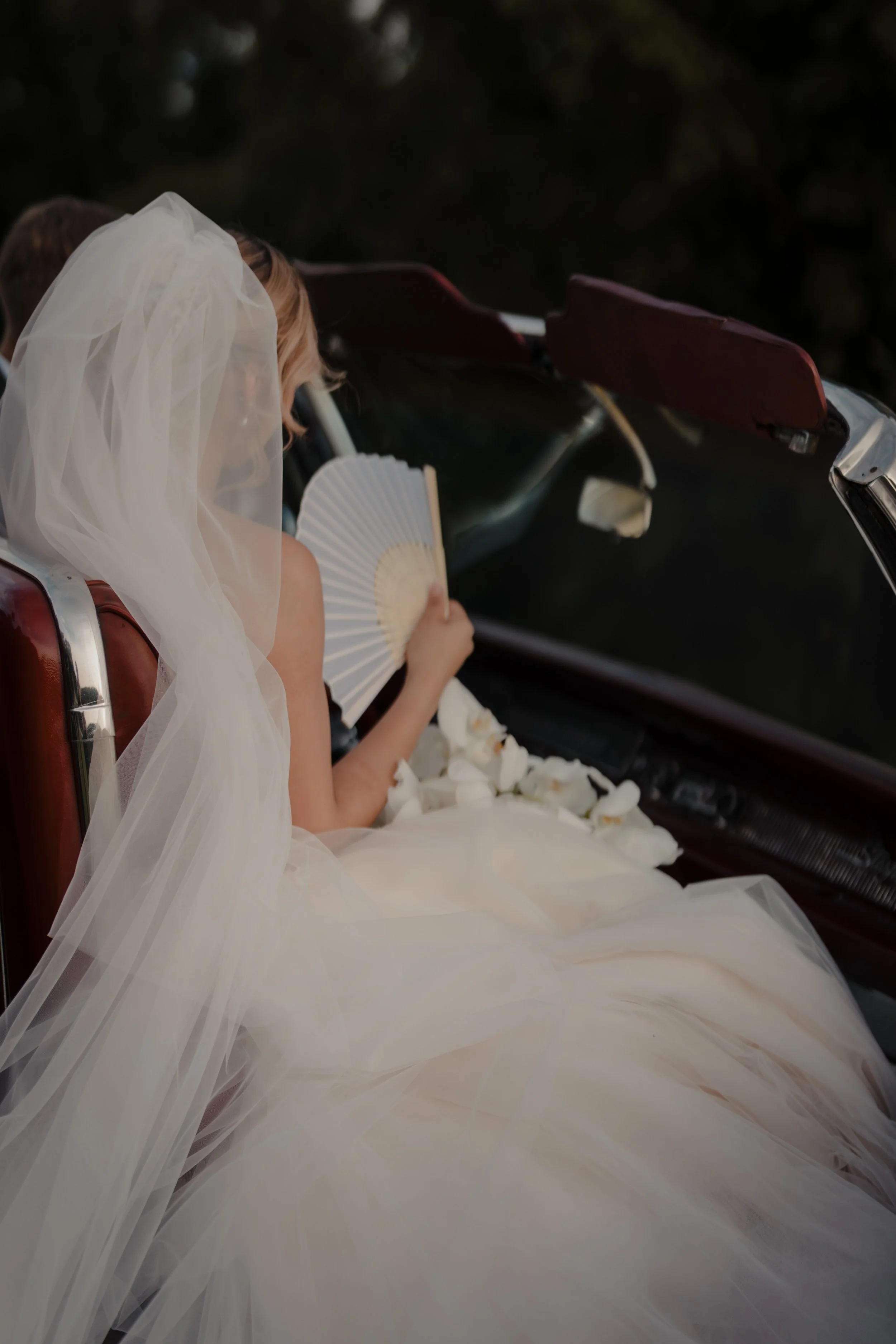 Mujer vestida de novia con velo y vestido de novia, sentada en un auto clásico, sosteniendo un abanico y rodeada de pétalos blancos.