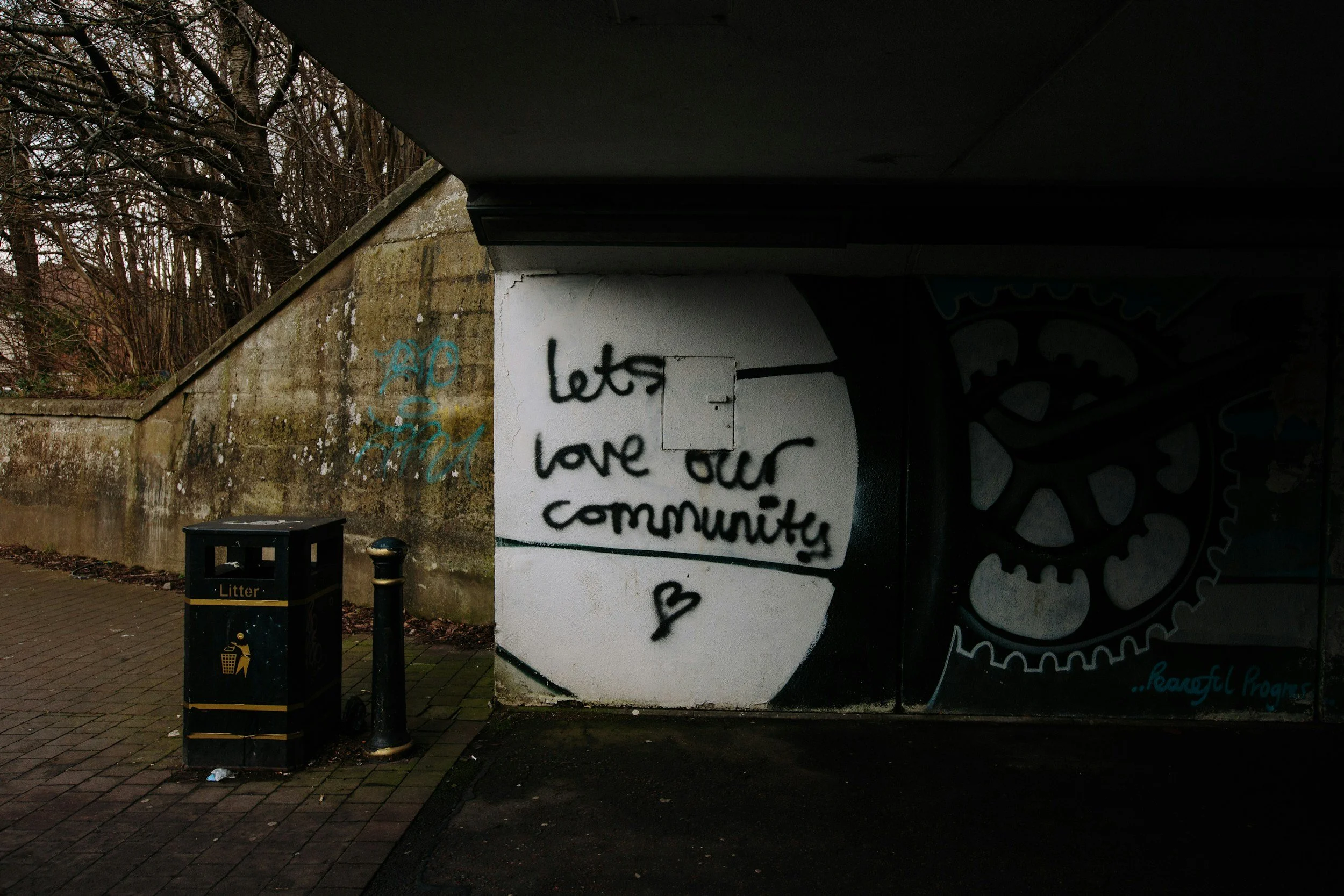 Graffiti on a white wall reading 'let's love our community' with a small heart, under a bridge with a support column, near a black litter bin and trash. Leafless trees are visible in the background.