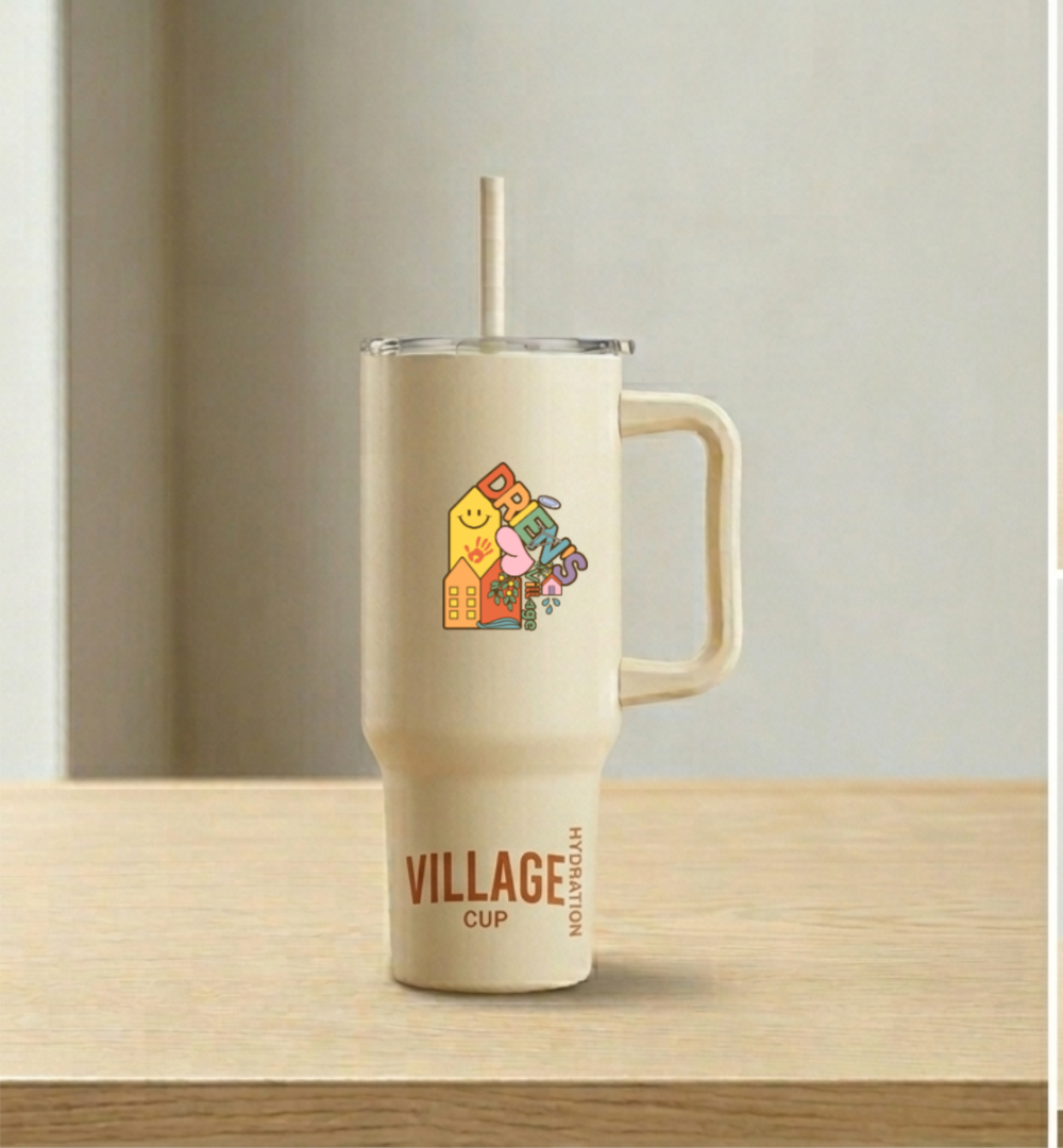 A beige travel mug with a straw, placed on a wooden surface. The mug has a sticker that says "DRIEN'S VILLAGE" with colorful graphics. The bottom of the mug features the words "VILLAGE CUP" and "HYDRATION".