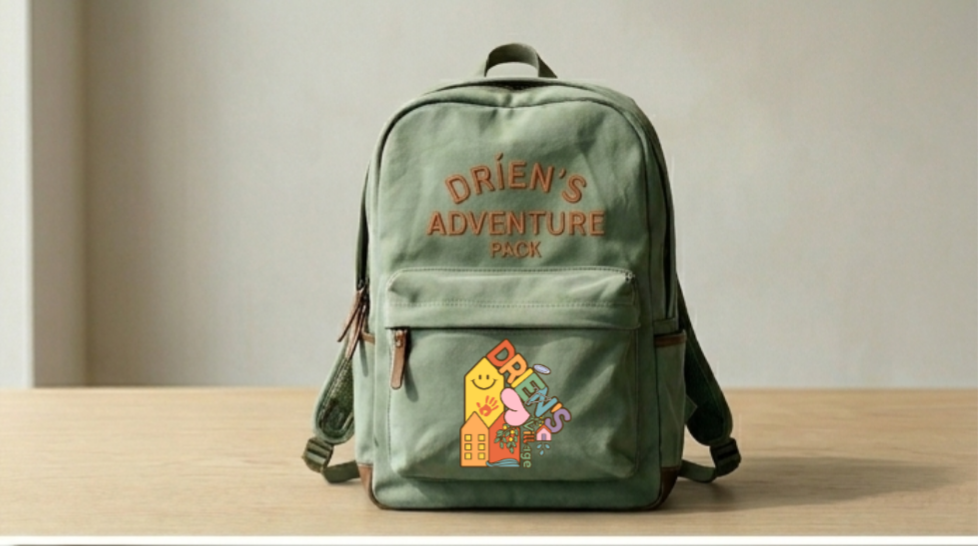 A canvas sustainable backpack with the words "DRIEN'S ADVENTURE PACK" embroidered on it, sitting on a wooden surface against a plain wall. A colorful sticker on the front with a smiling yellow character and other playful designs.