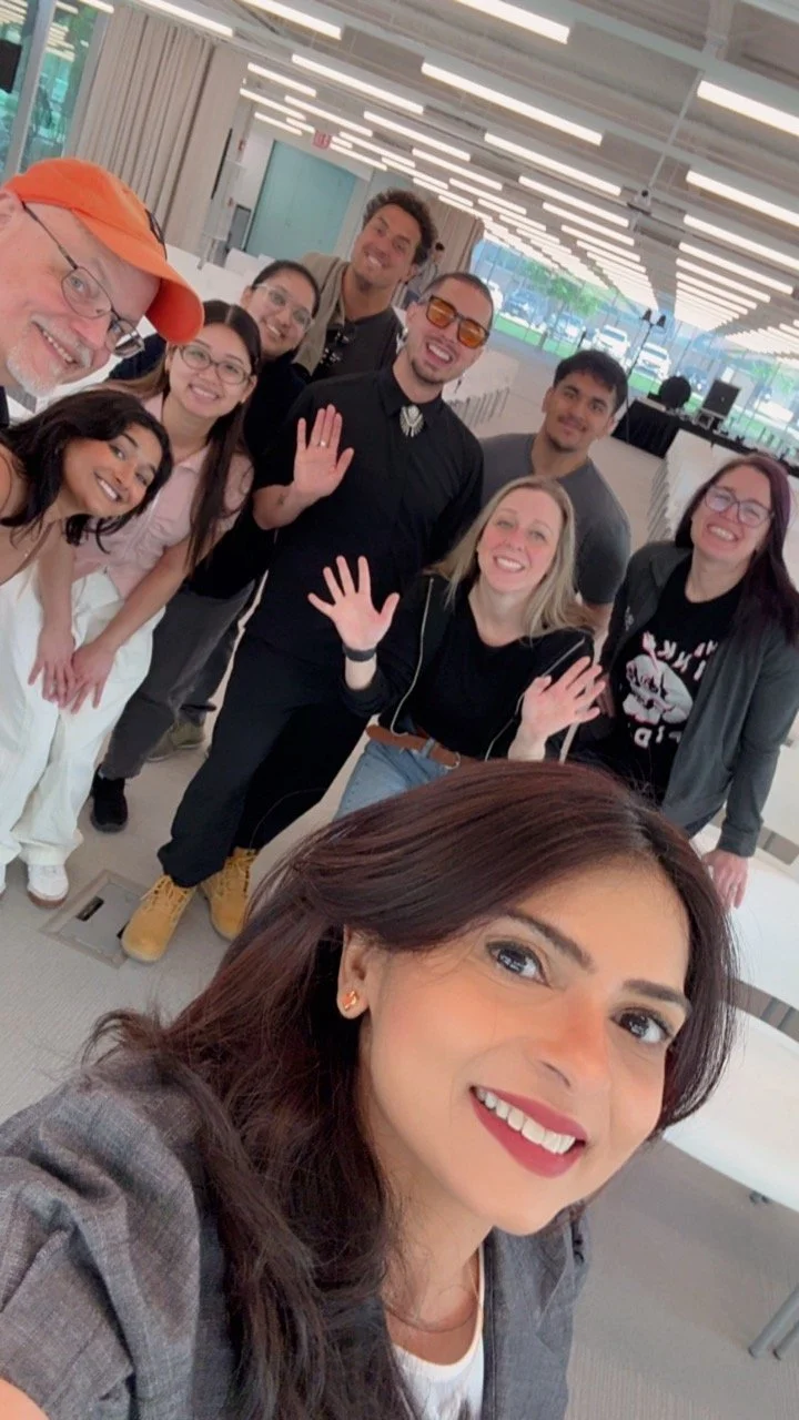 Group of smiling people in an indoor setting, taking a selfie with a woman in the foreground, making peace signs and waving.