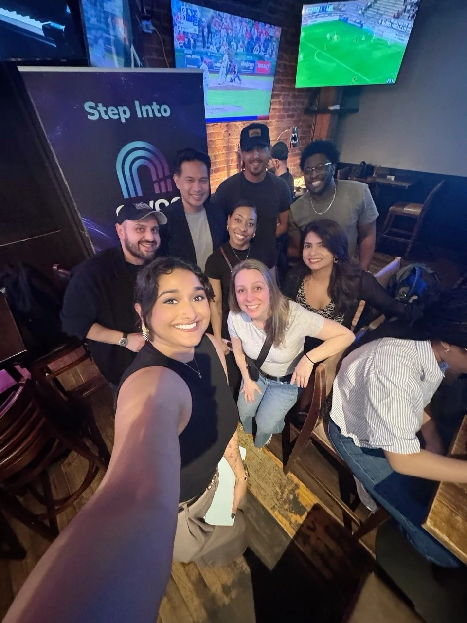 A group of nine diverse young adults smiling and posing for a selfie in a bar or restaurant with large TVs showing sports games in the background.