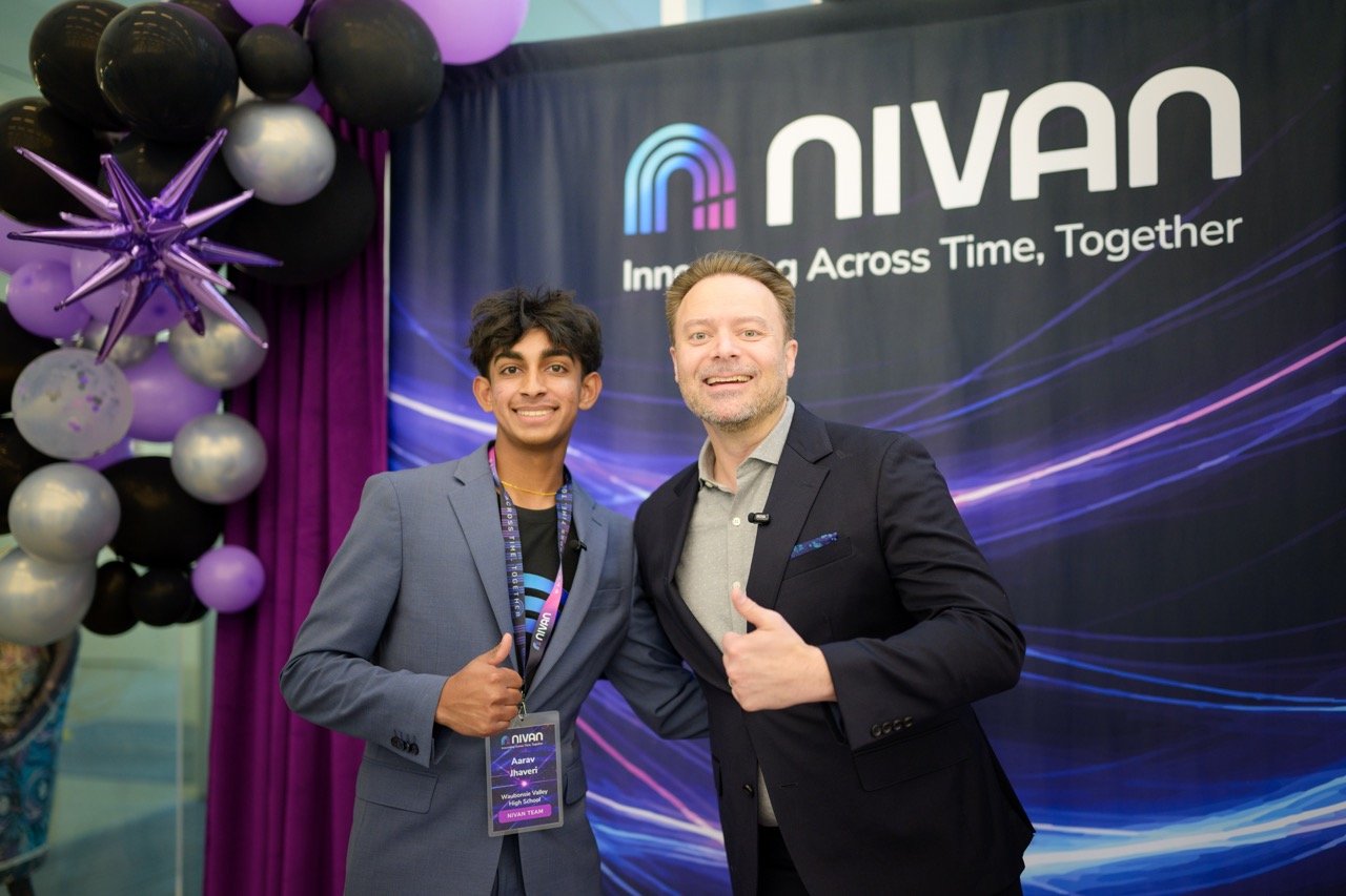 Two men in suits standing in front of a NIVAN banner at an event, with balloons decorated in purple, black, and silver.