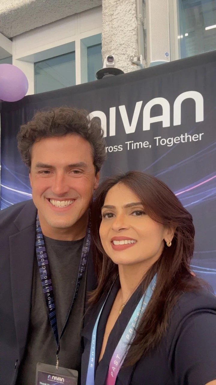 Two people taking a selfie in front of a black banner with the word "NIVAN" and the slogan "Cross Time, Together." The man has curly hair and is smiling, wearing a dark blazer and a conference badge. The woman has long brown hair and is also smiling,