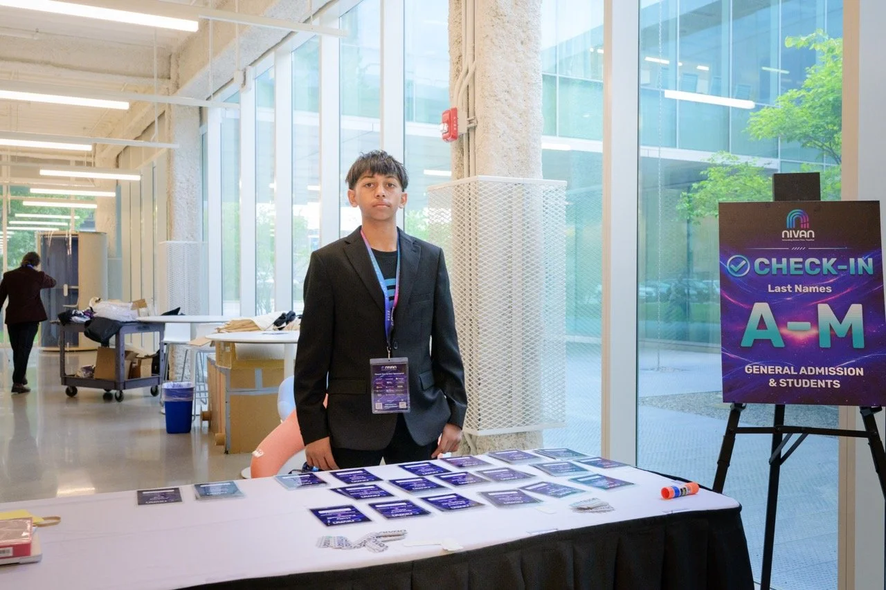 A young man in a black blazer standing at a registration table with check-in materials, inside a modern building with large windows, a registration sign with last names A-M, and a person in the background.