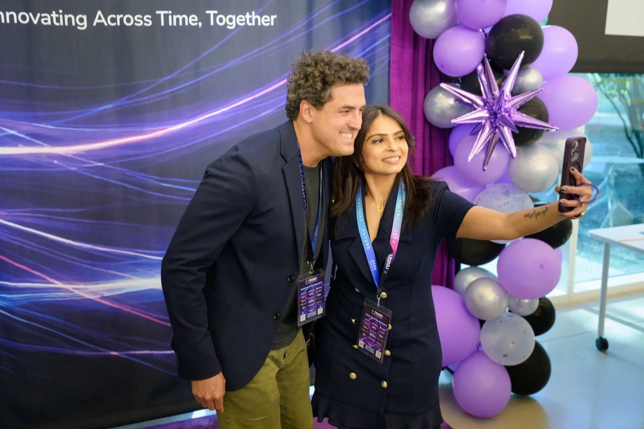A man and a woman taking a selfie together at a conference or event. The woman is holding a smartphone and both are smiling. They are standing in front of purple and black balloons with a decorative star-shaped balloon. The background features a dark