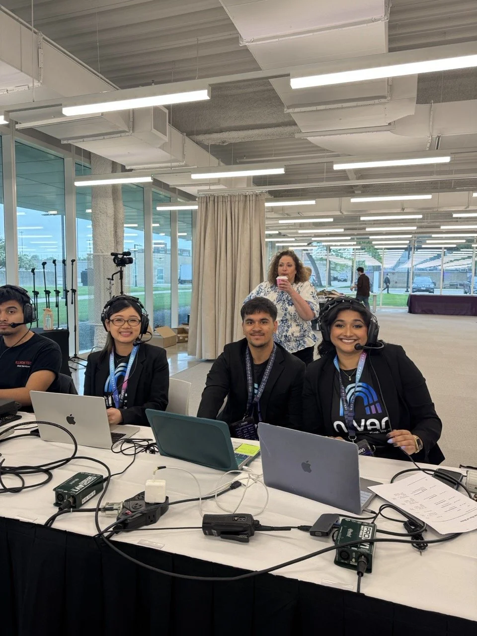 Four people wearing headsets and sitting at a table with laptops, with a woman standing behind them in a bright indoor space with large windows and ceiling lights.