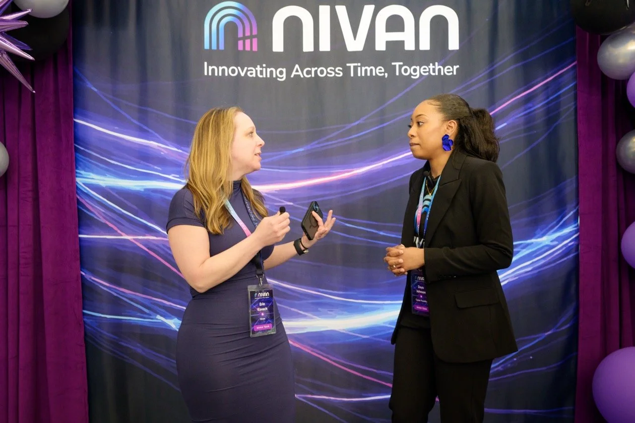 Two women speaking at a Nivan conference, with a backdrop displaying the Nivan logo and the slogan 'Innovating Across Time, Together,' surrounded by purple curtains and balloons.