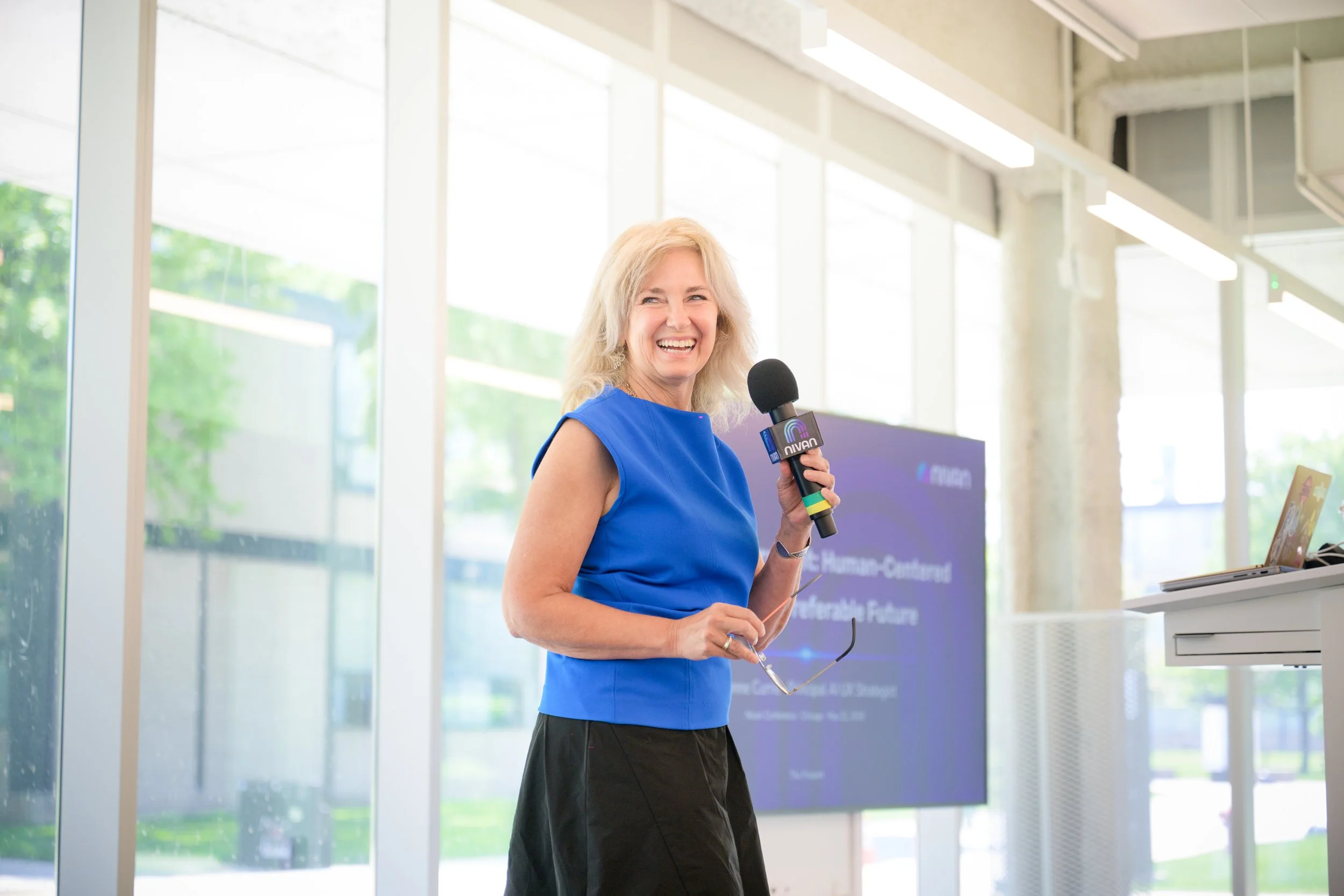 A woman with blonde hair in a blue sleeveless dress smiling while holding a microphone in front of a large window during a presentation or event.