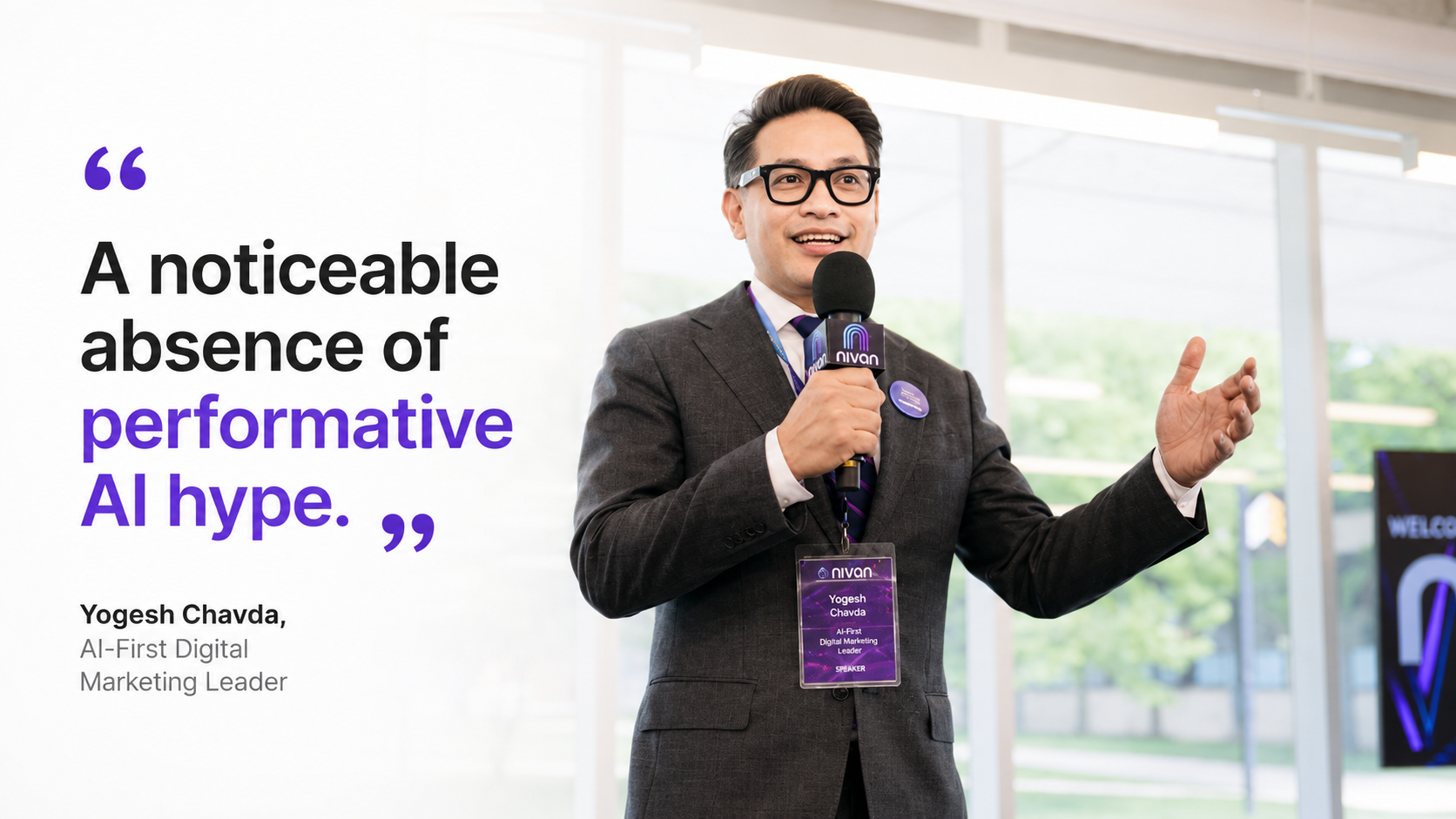 Yogesh Chavda, AI-First Digital Marketing Leader, speaking tat Nivan Live with a microphone in hand, in a modern indoor setting with large windows and a screen displaying a welcoming message in the background.