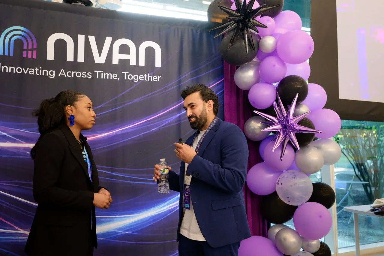 A man and a woman are having a conversation at a conference or event. The woman is wearing a black blazer and blue earrings. The man is dressed in a blue blazer, holding a water bottle, and has a lanyard with a badge around his neck. They are standin