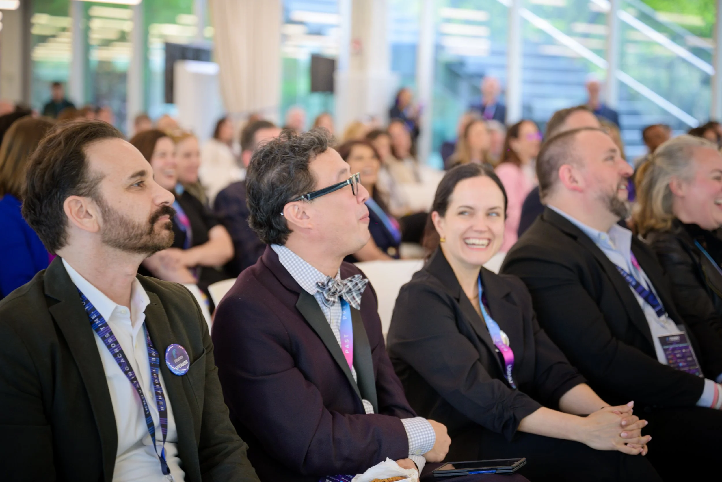Audience attending a conference or seminar, sitting in rows, facing forward, some smiling, in a bright room with large windows.