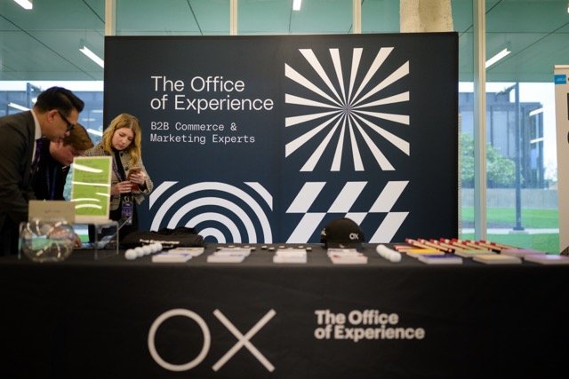A large sign reading 'The Office of Experience B2B Commerce & Marketing Experts' with geometric black and white patterns in a modern conference setting. Two people are looking at a table with various items near the sign.
