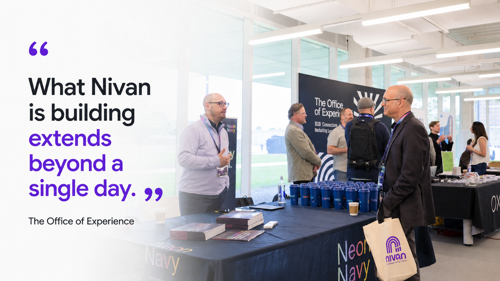 A professional at Nivan Live or expo with a booth for Nivan, featuring banners that read 'The Office of Experience'. Several people are engaging in conversations; a man with glasses and a dark jacket is holding a tote bag with the Nivan logo. 