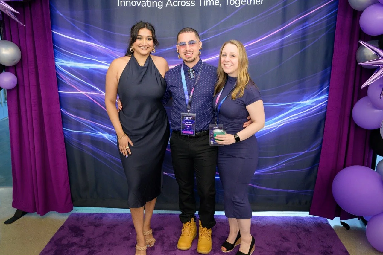 Three people standing together at an event, smiling. They are in front of a backdrop with blue and purple light streaks, with purple curtains and balloons around them. The person on the left is a woman in a navy dress, the person in the middle is a m